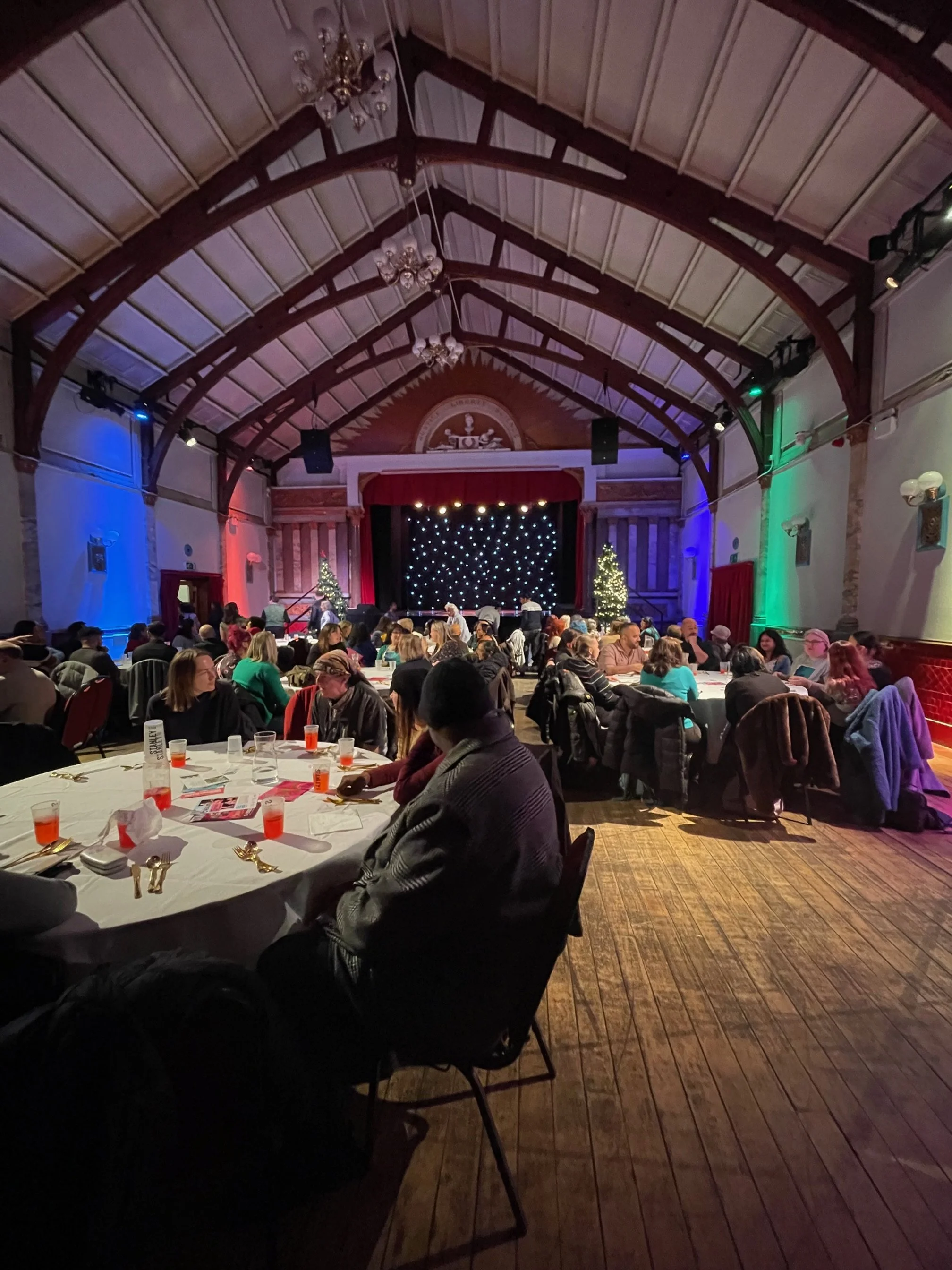 A large gathering of people attending an event in a decorated hall with a stage, Christmas trees, and string lights, seen from the perspective of seated guests at round tables.