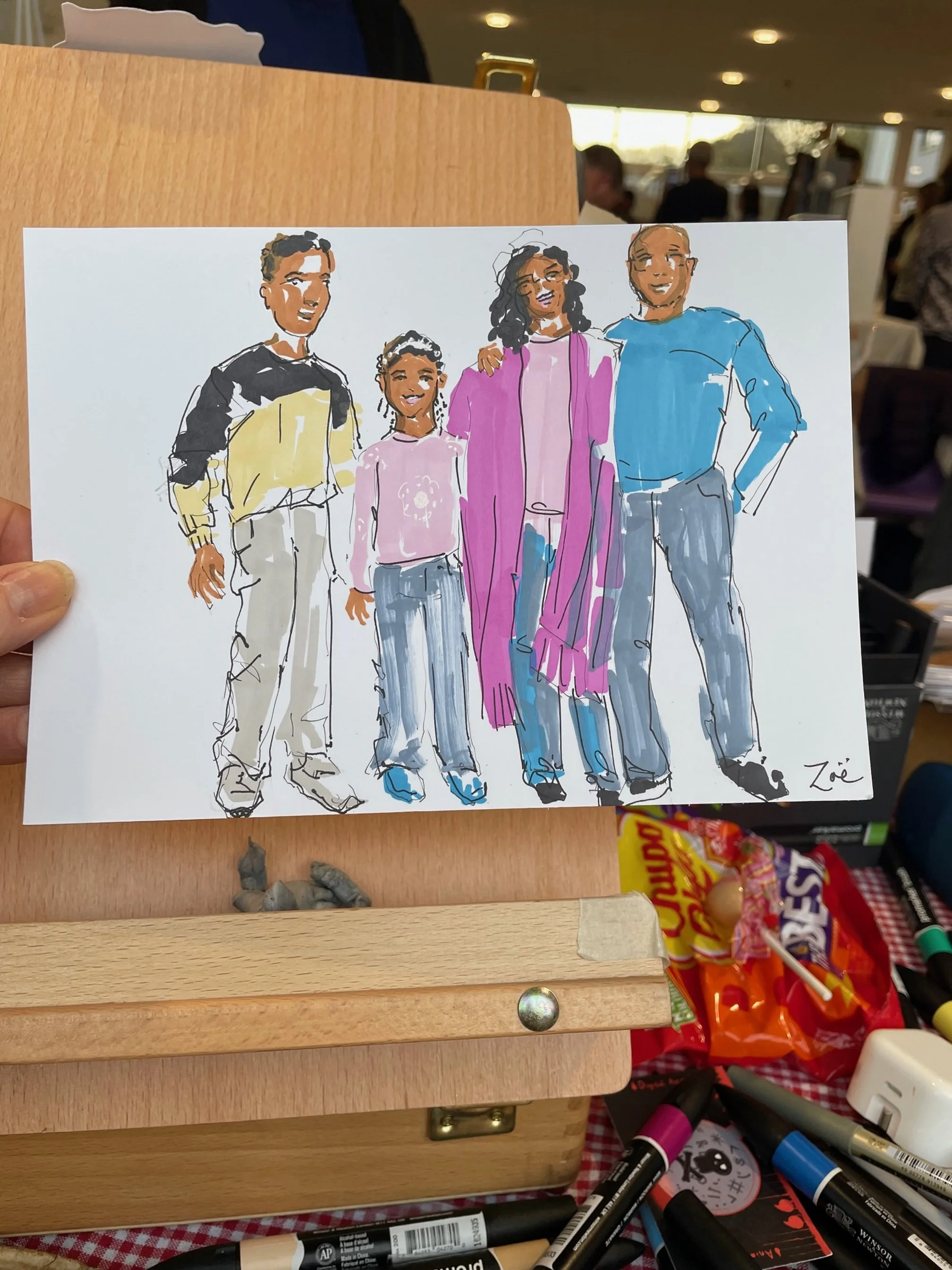 Hand-drawn illustration of a family of five standing together in a line, smiling. The family includes two men, two women, and a young girl. The background shows an indoor setting with blurred people and objects.