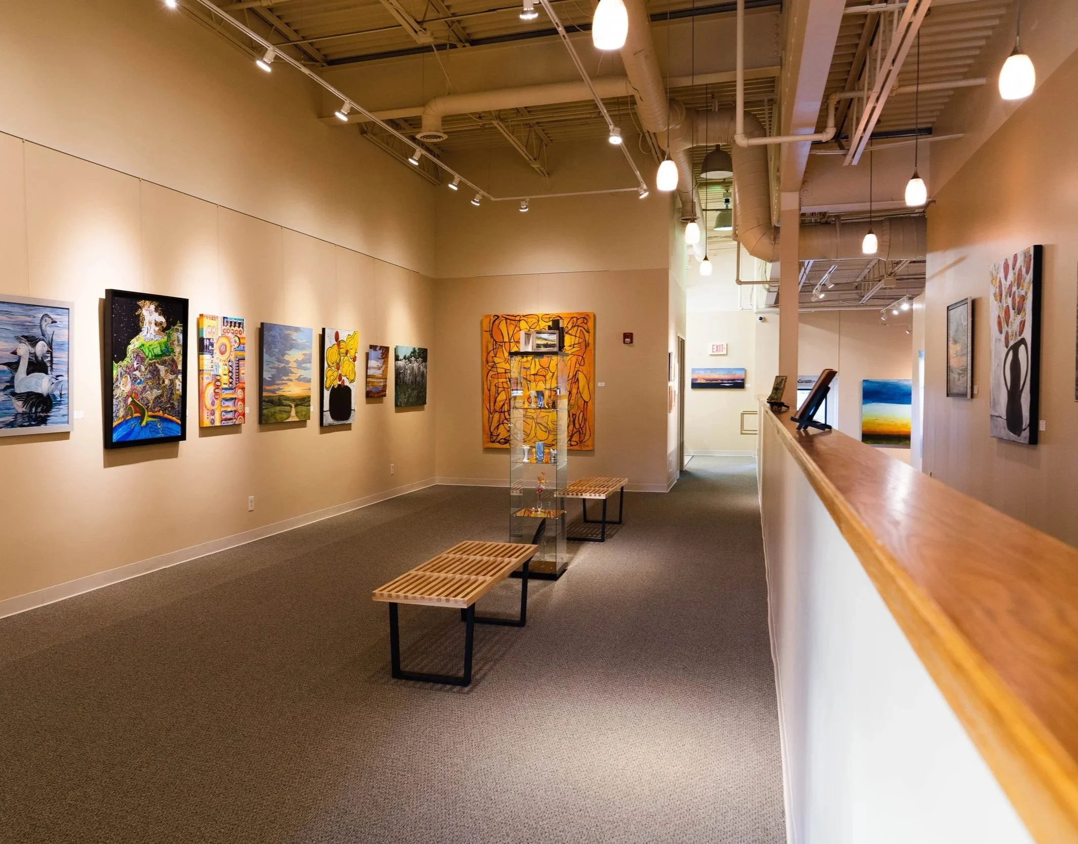 Interior of an art gallery with various paintings on beige walls, benches in the foreground, and track lighting on the ceiling.