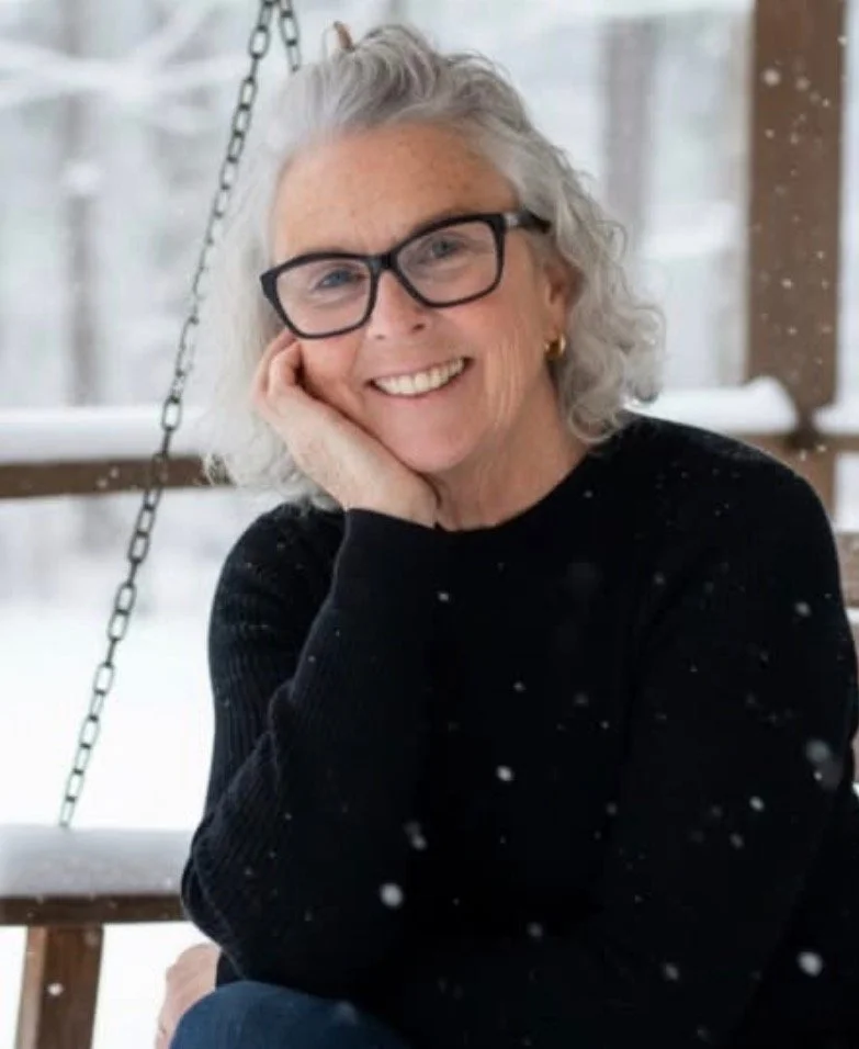 An elderly woman with gray hair, glasses, and a black sweater smiling while sitting outdoors in snowy weather.