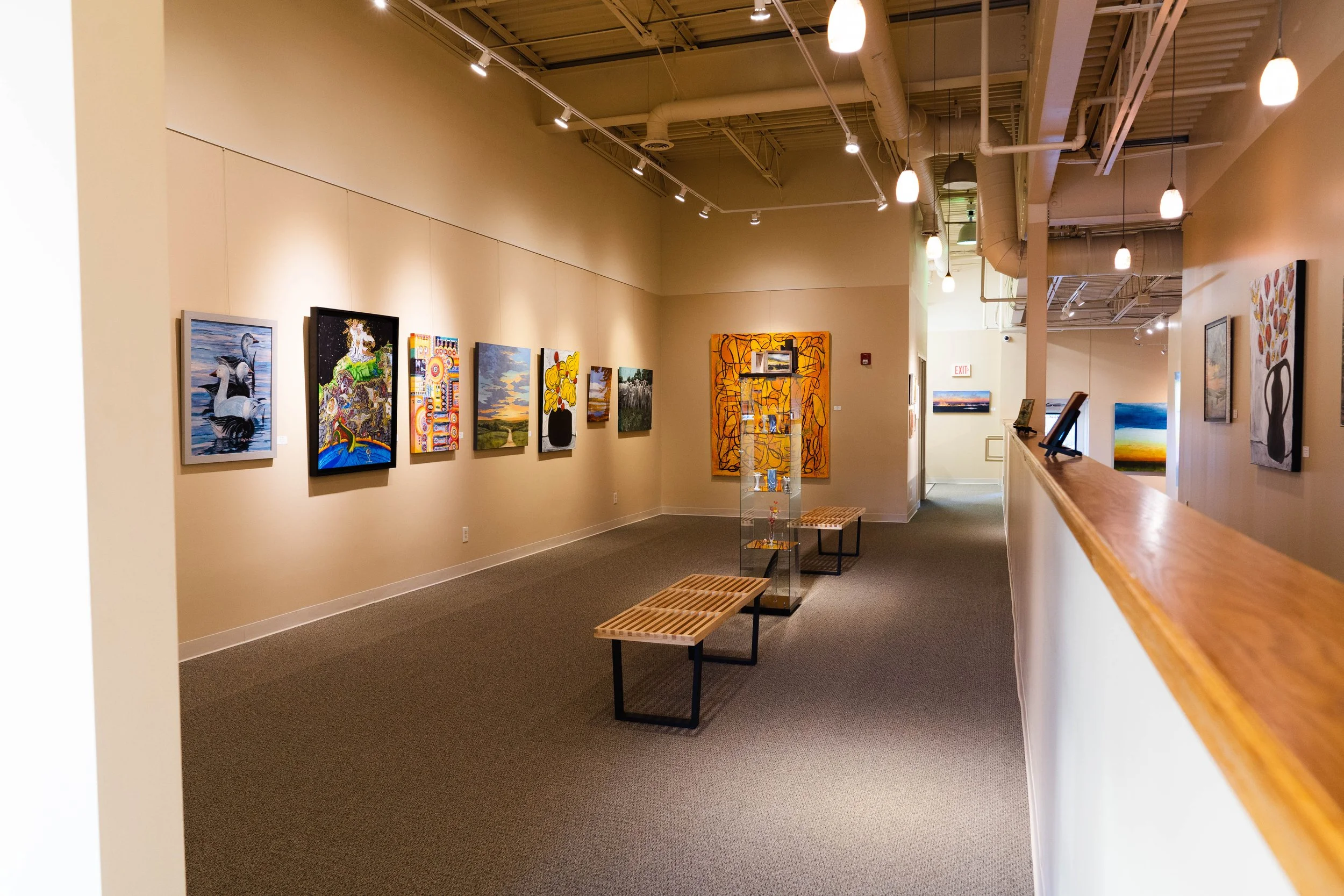 Interior of The Fusion art gallery at Parkville Frame Gallery with paintings on beige walls, track lighting, benches, and a glass display case in the center.