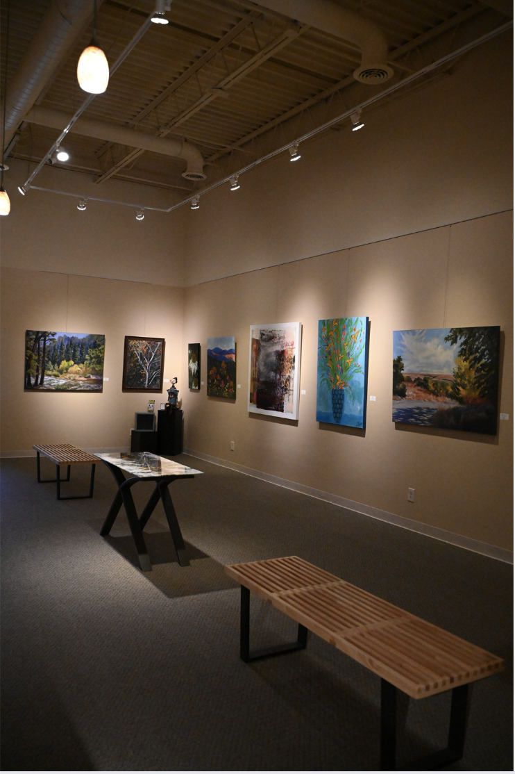 Interior of The Fusion at Parkville Frame Gallery - Interior of an art gallery with several landscape paintings on beige walls, wooden benches, and a table with a decorative glass piece, overhead lighting illuminating the artwork.