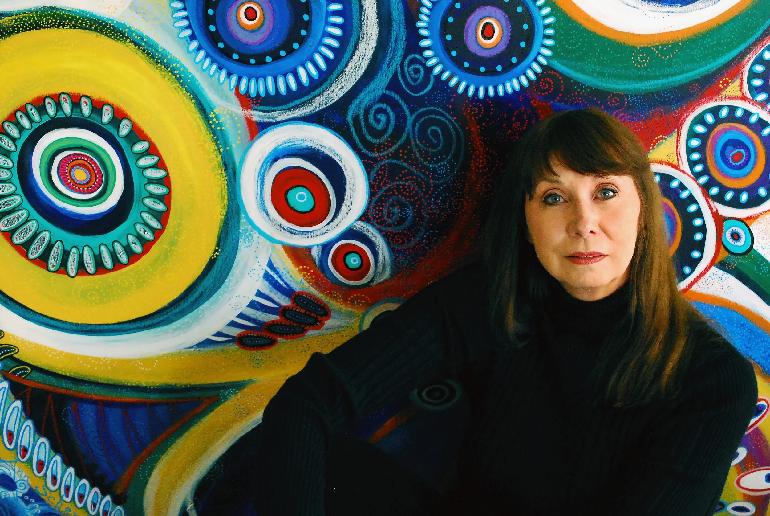 A woman with long brown hair and blue eyes sitting in front of a colorful abstract mural with circular patterns and vibrant colors.