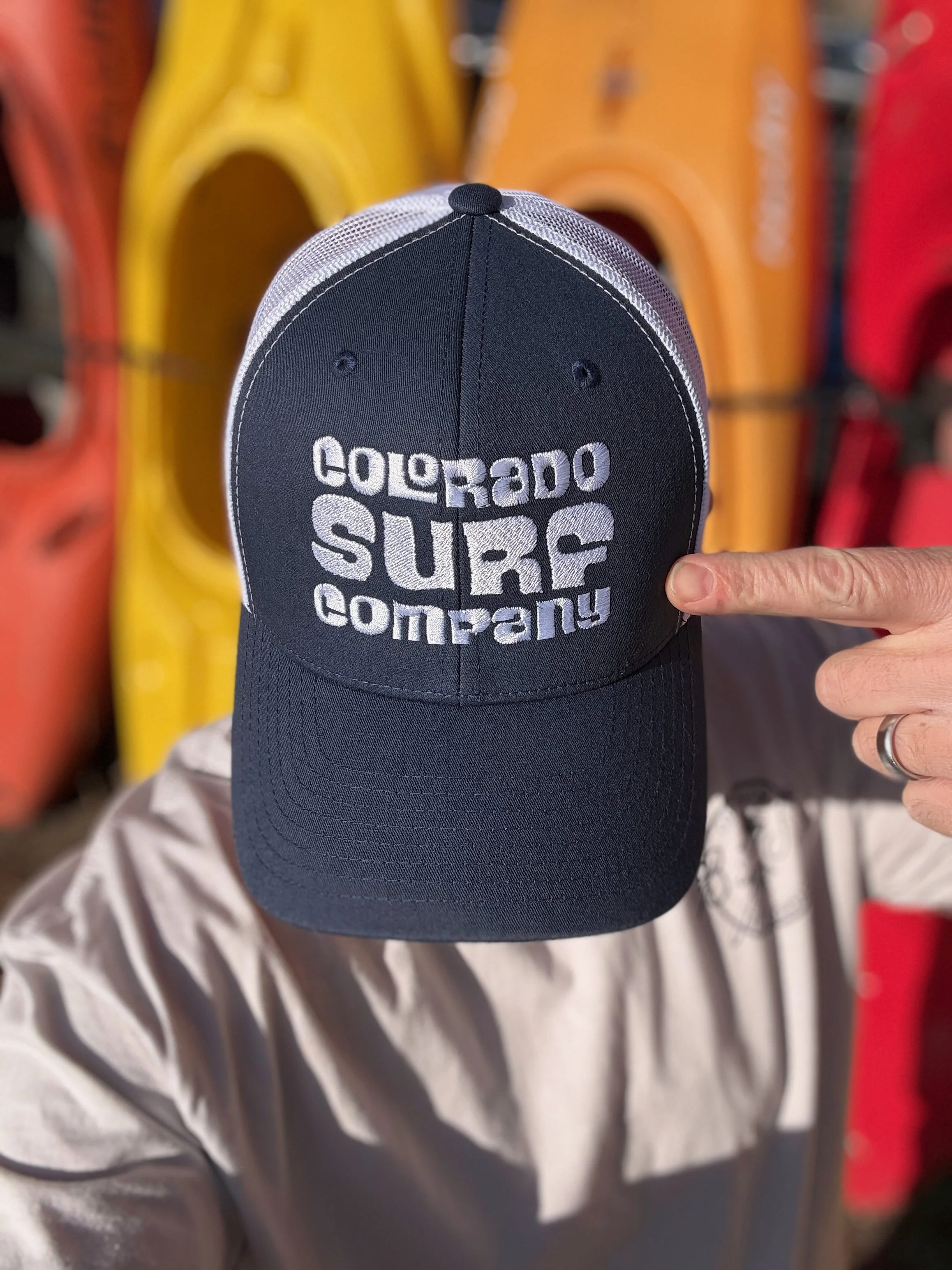 A person's hand pointing at a black baseball cap with white embroidery that reads 'COLORADO SURF COMPANY,' held in front of a display of kayaks in various colors.