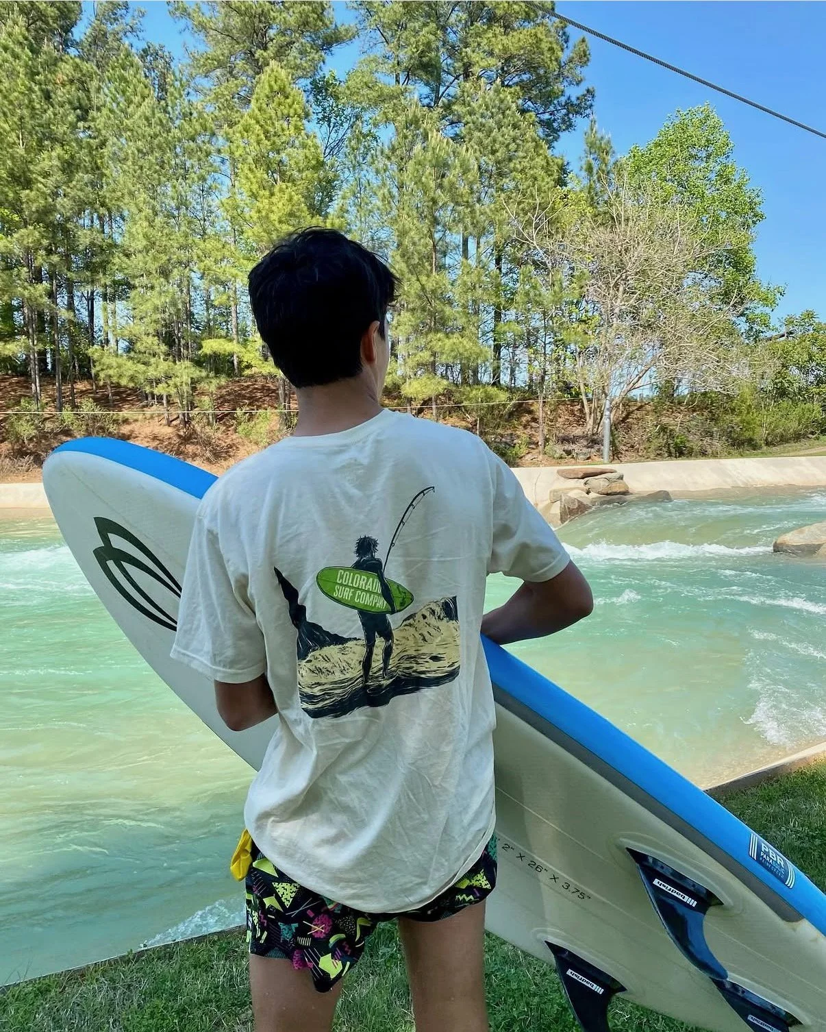 A boy holding a paddleboard at a water park or river, wearing a beige t-shirt with a surfer graphic on the back, and colorful shorts, standing near water with trees and rocks in the background.