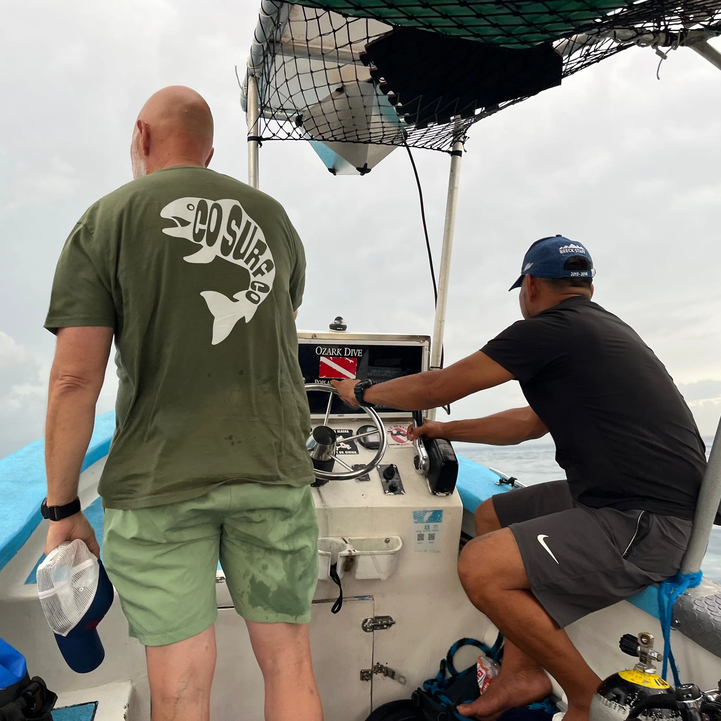 Multiple people are working on repairing or maintaining equipment on a boat, with one person kneeling and others standing around, under a cloudy sky.