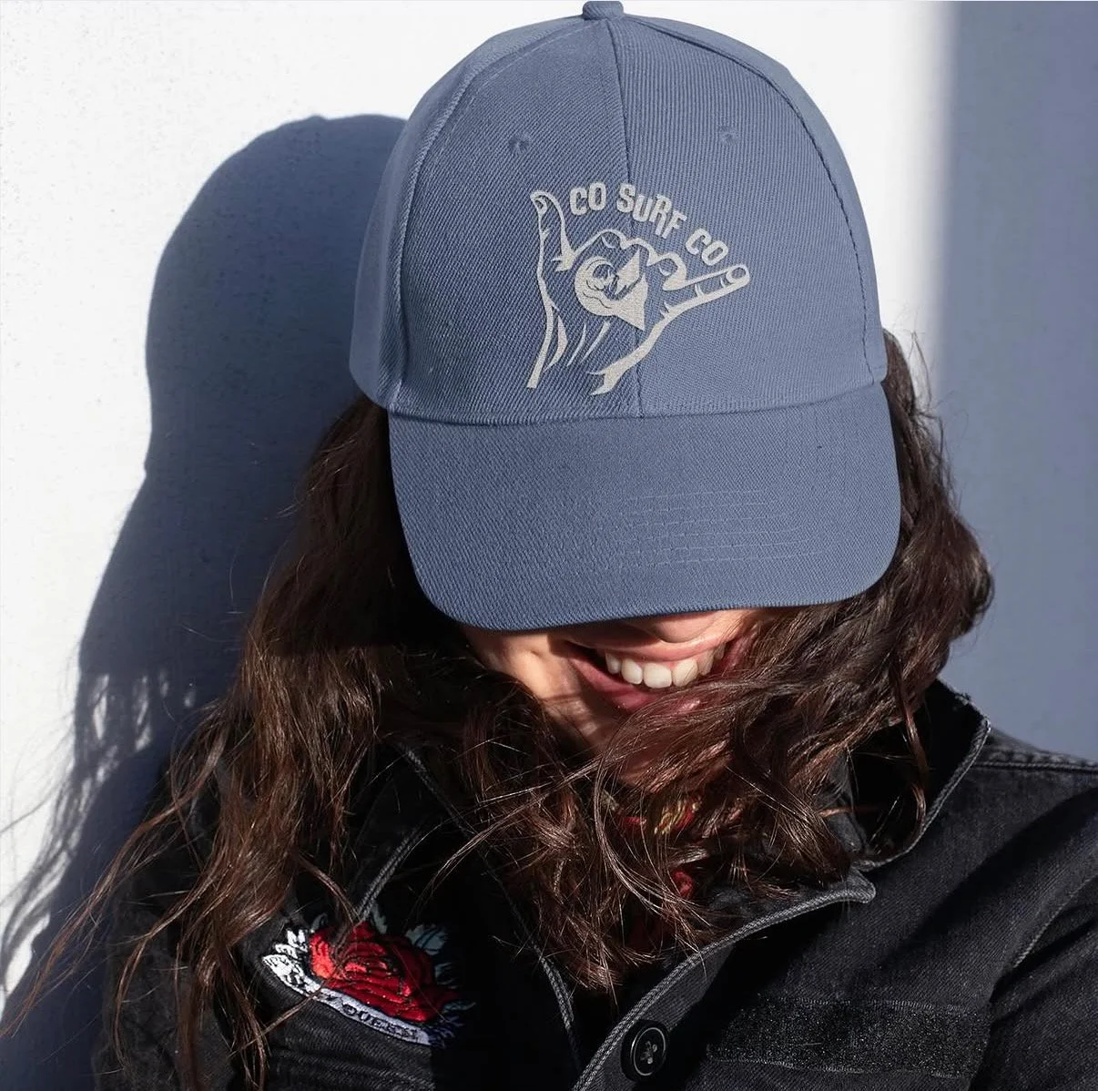 Person wearing a gray cap with a griffin logo and the words 'CO SURF CO 9', smiling with curly brown hair, partially covered by the cap. Wearing a black jacket with a rose embroidery patch.