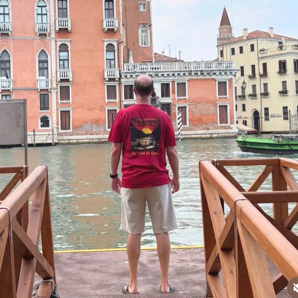 A man standing on a dock facing a canal with colorful historic buildings across in Venice, Italy, during daytime.