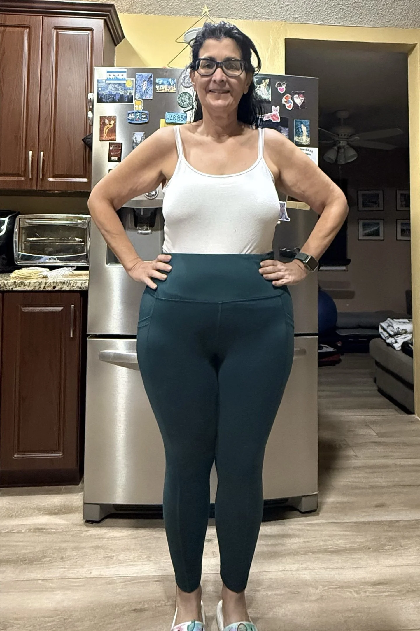 A middle-aged woman with dark hair and glasses stands in a kitchen, smiling. She is wearing a white tank top, teal high-waisted leggings, and white shoes. She has her hands on her hips.