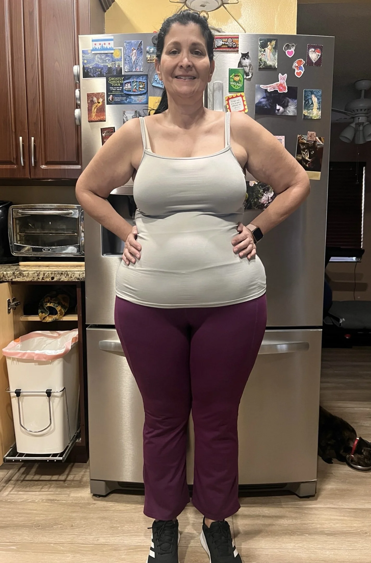 A woman stands in a kitchen with her hands on her hips, wearing a light gray tank top and maroon pants, with a black smartwatch on her left wrist. The refrigerator behind her has various magnets and pictures, including animals and scenic views. To her right, a black and brown dog is lying on the wooden floor.