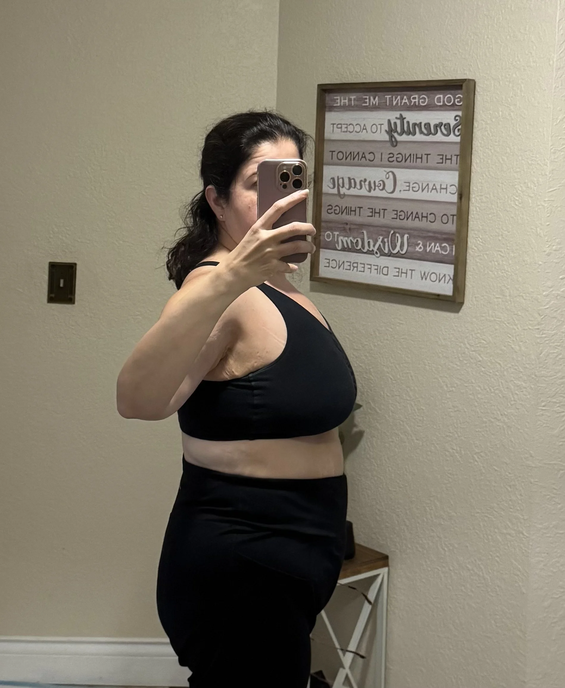 Side view of a woman taking a mirror selfie in a black sports bra and black pants, with a framed sign on the wall behind her.