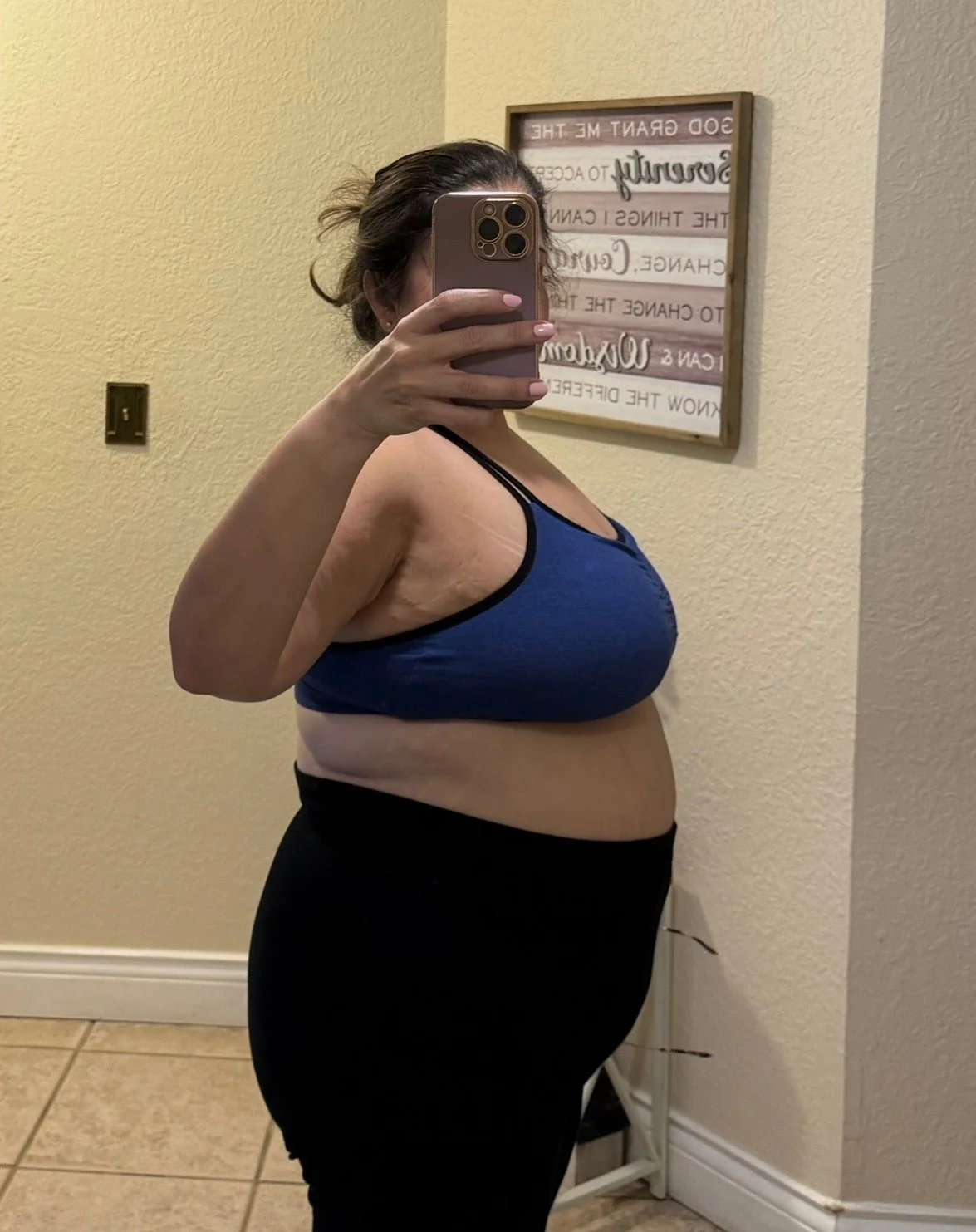 A woman taking a mirror selfie with her smartphone, showing her side profile in a room with beige walls and a framed quote on the wall.