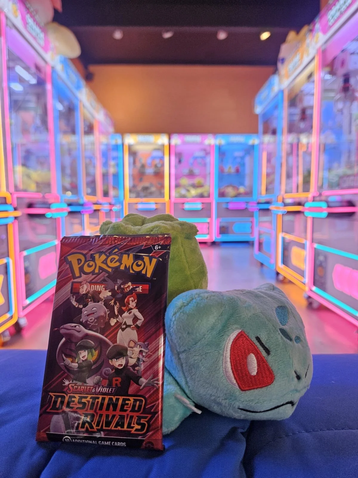 A Pokémon card pack and two plush toys of Bulbasaur and Squirtle in a colorful arcade with neon lights and claw machines in the background.