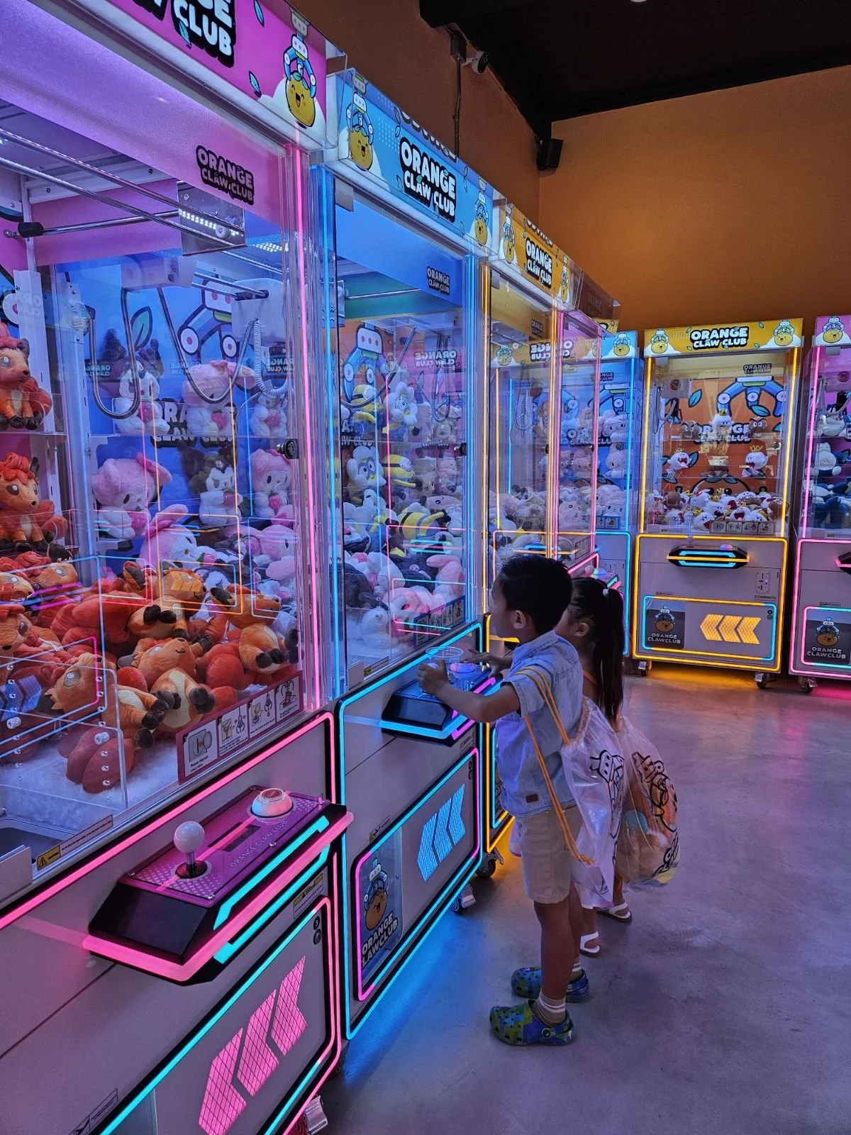 Children playing arcade claw machine game at an entertainment center with neon lighting and plush toys inside the machine.