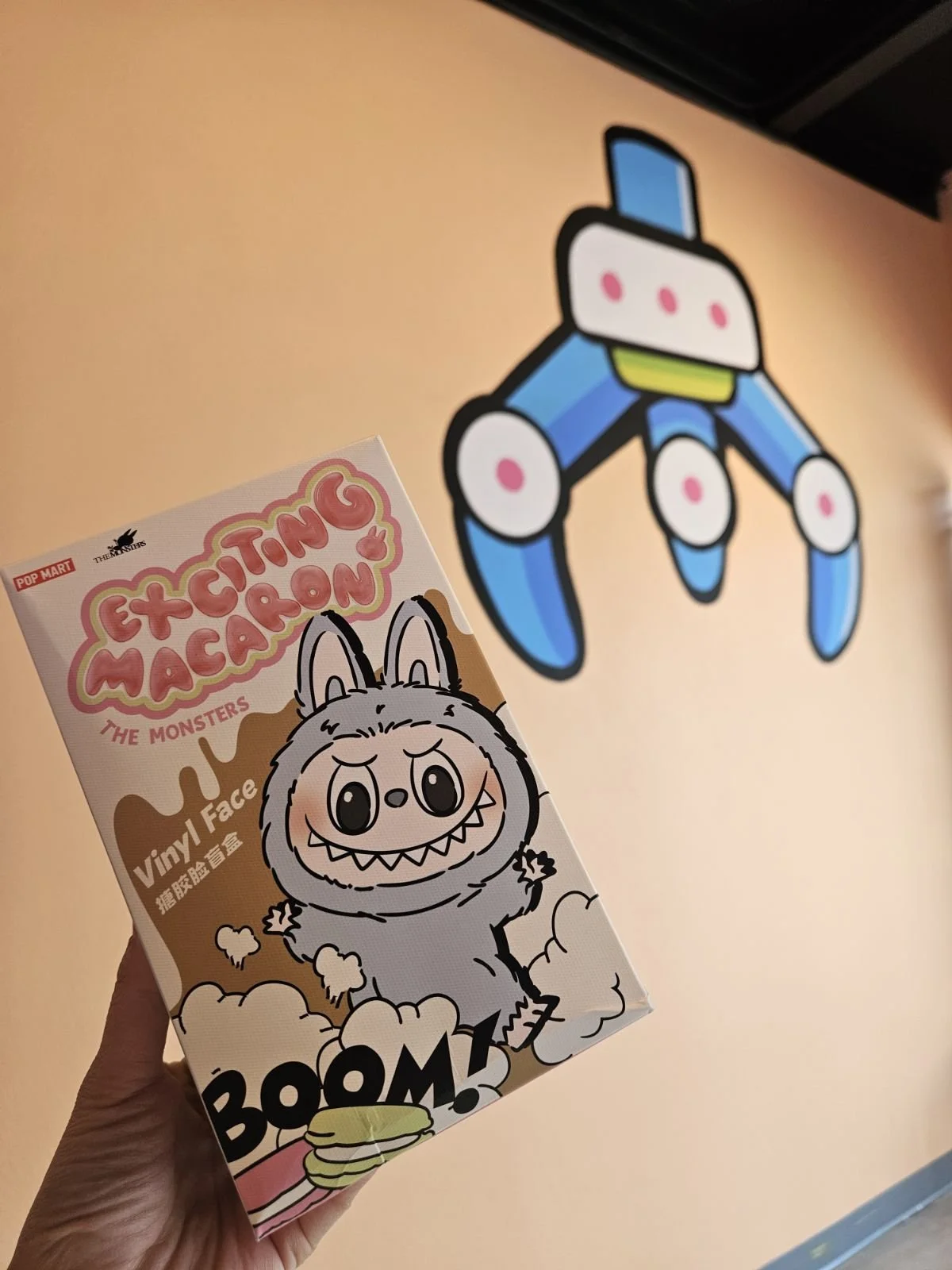 Person holding a box of Extingu M-shaped vinyl face mask with a cartoon monster illustration held in front of the Orange Claw Club mural in Downtown Santa Ana.