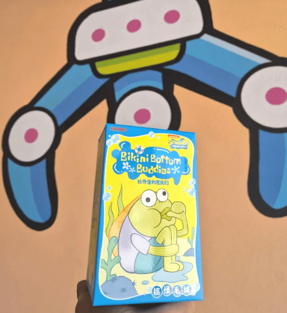 An image of a Spongebob Bikini Bottom popmart Blind Box being held in front of the Orange Claw Club mural in Santa Ana.