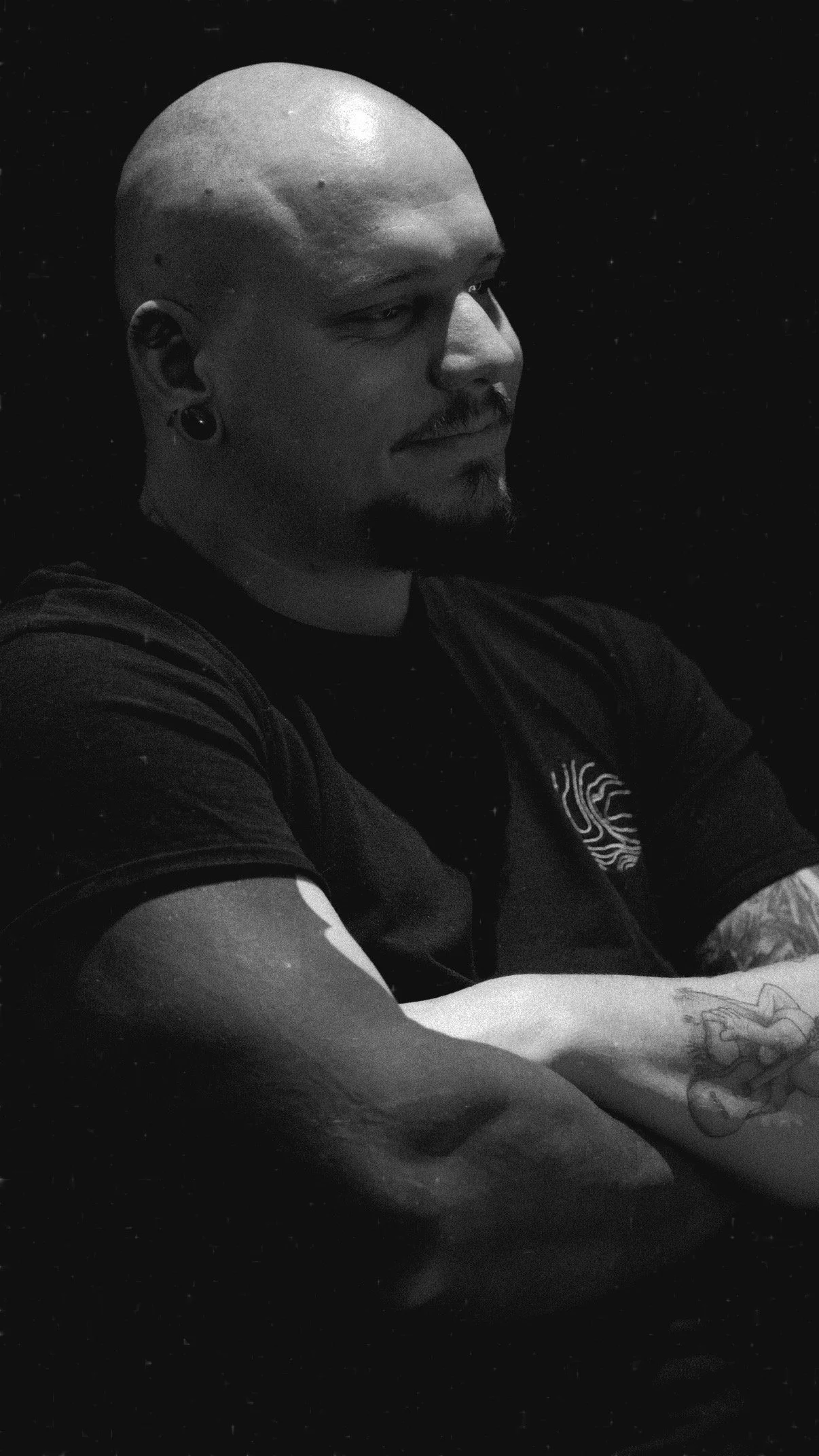 Portrait en noir et blanc de Wootz, tatoueur, les bras croisés et le regard tourné vers la droite.