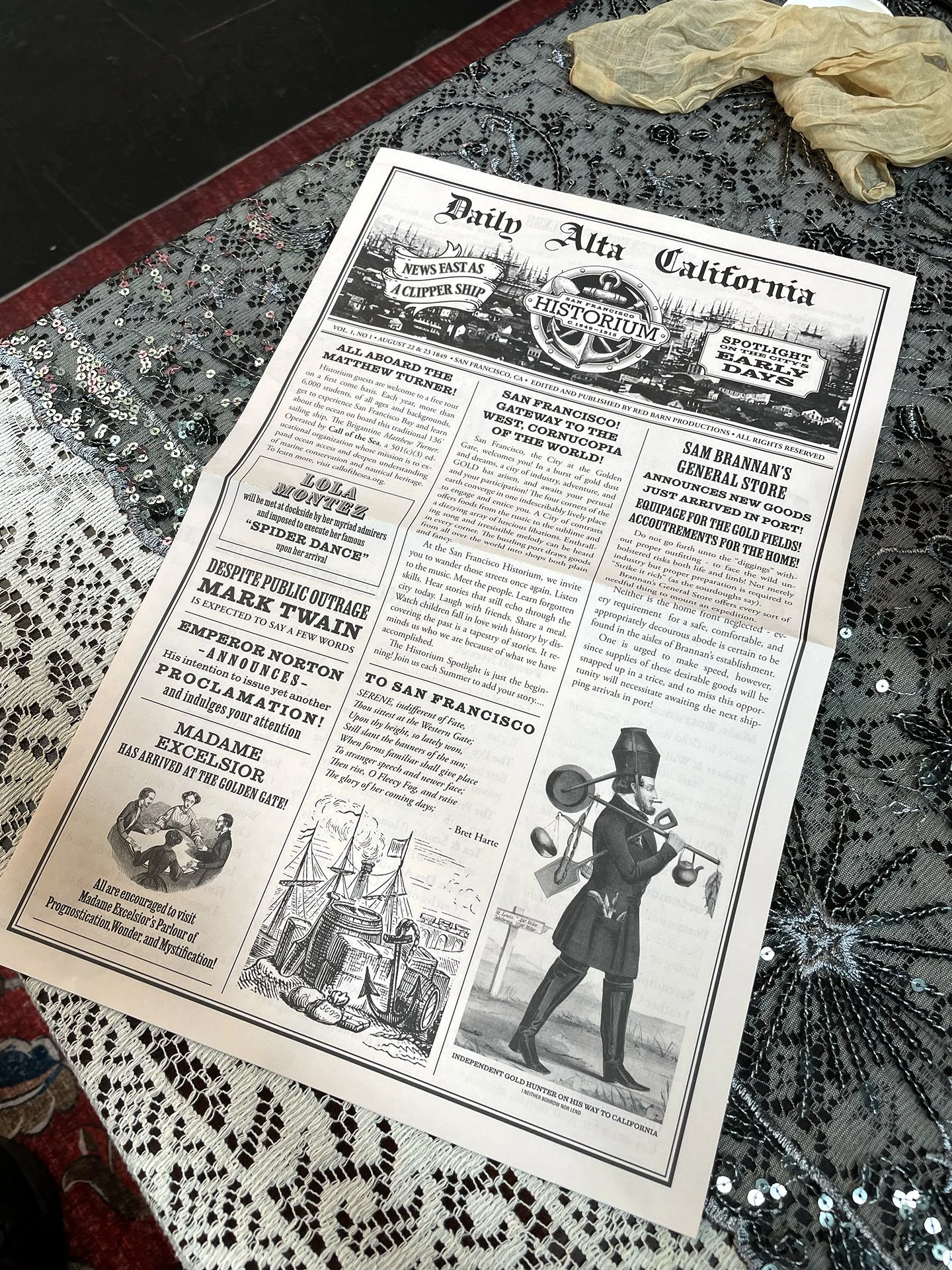 Event program mimicking a vintage-style newspaper titled "Daily Alta California" with illustrations of ships and a gold miner, resting on a textured, sequined fabric.