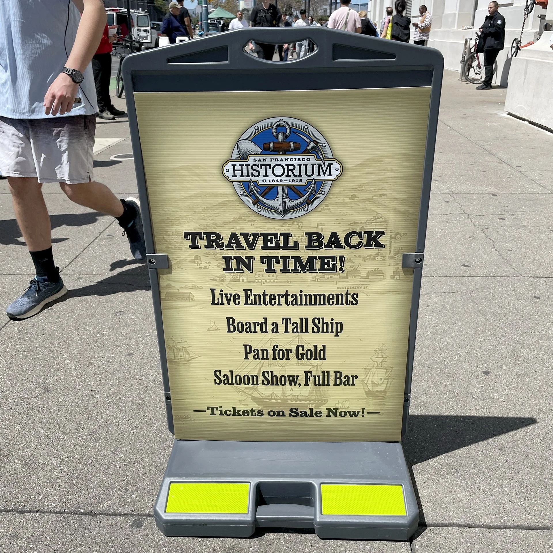 A signboard advertising an event at the San Francisco Historicum with activities like live entertainment, boarding a tall ship, pan for gold, saloon show, and full bar. The sign is placed outdoors on a sidewalk with people walking in the background.