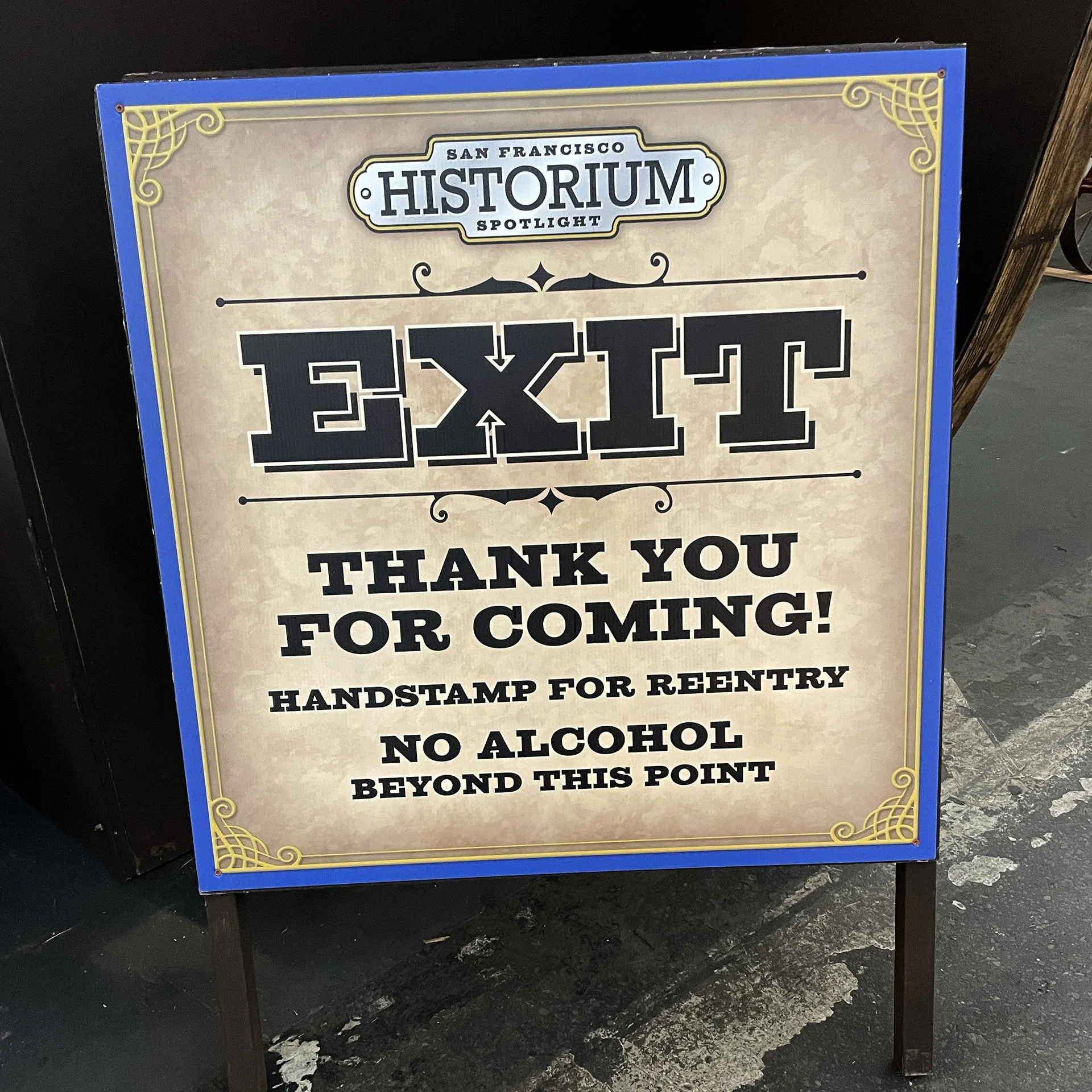 A sign with a blue and gold border reading 'San Francisco Historium Spotlight' at the top, with large black text saying 'EXIT'. The sign also thanks visitors for coming, mentions handstamps for reentry, and states no alcohol beyond this point.