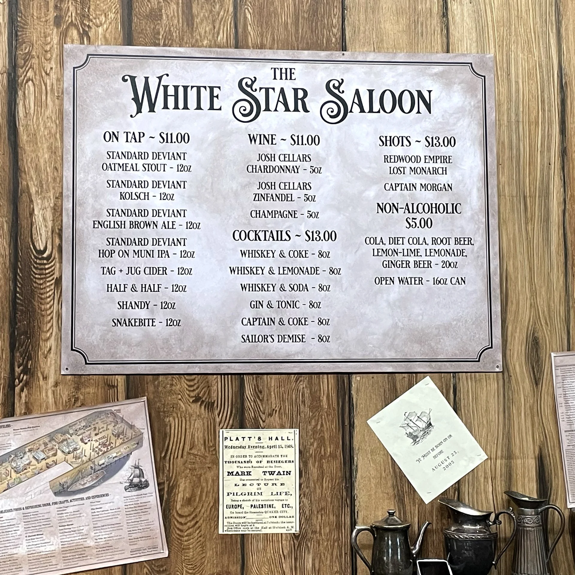 A wooden wall with a sign that reads 'The White Star Saloon' listing drinks, wine, cocktails, shots, and non-alcoholic beverage prices. Several historical documents and illustrations are pinned on the wall, along with a tea set.