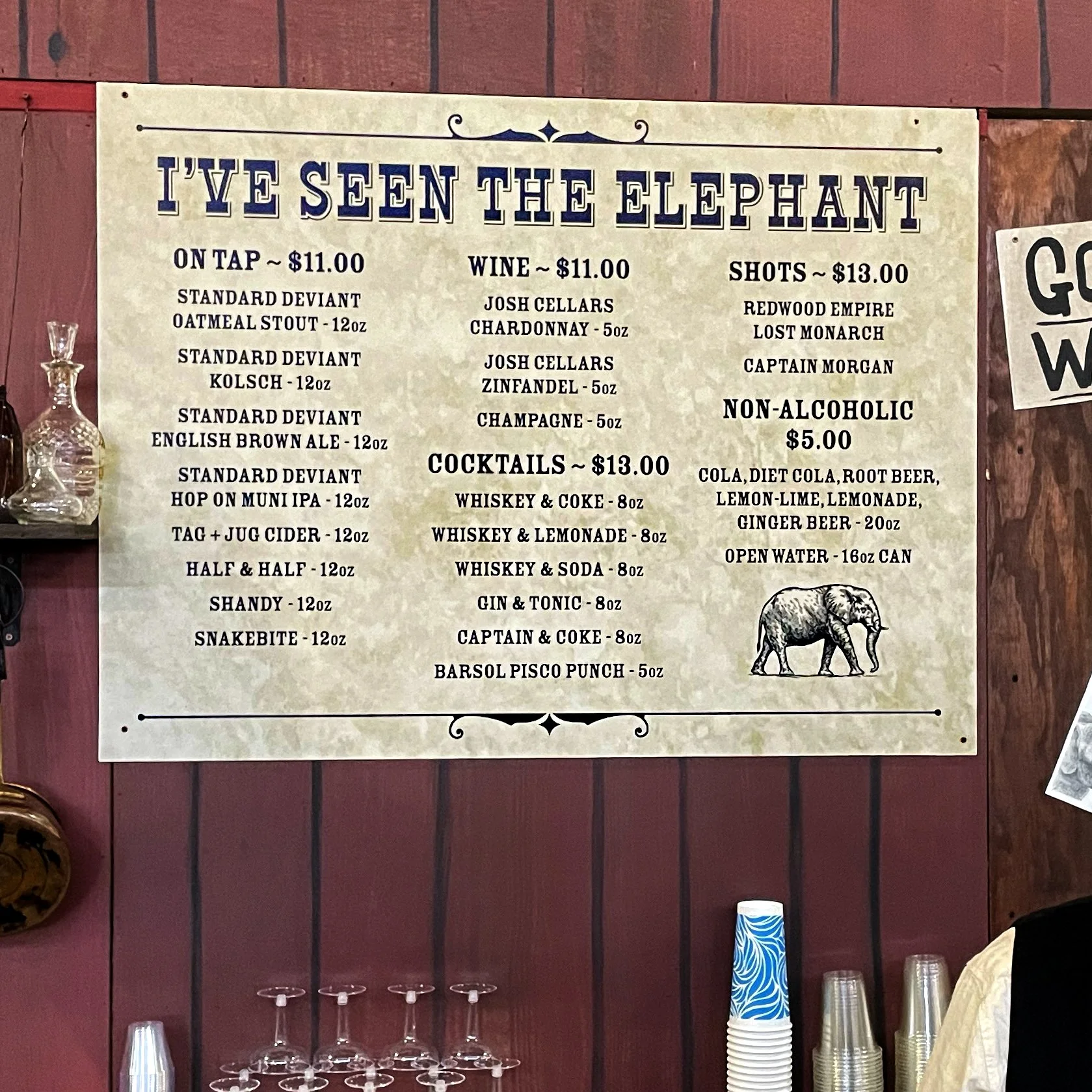 Menu board titled "I've Seen the Elephant" listing drinks and prices: beer on tap for $11, wine for $11, shots for $13, non-alcoholic options for $5, and cocktails for $13. The drinks include various beers, wines, and mixed drinks. Part of a bar setting with glasses and cups below the board.