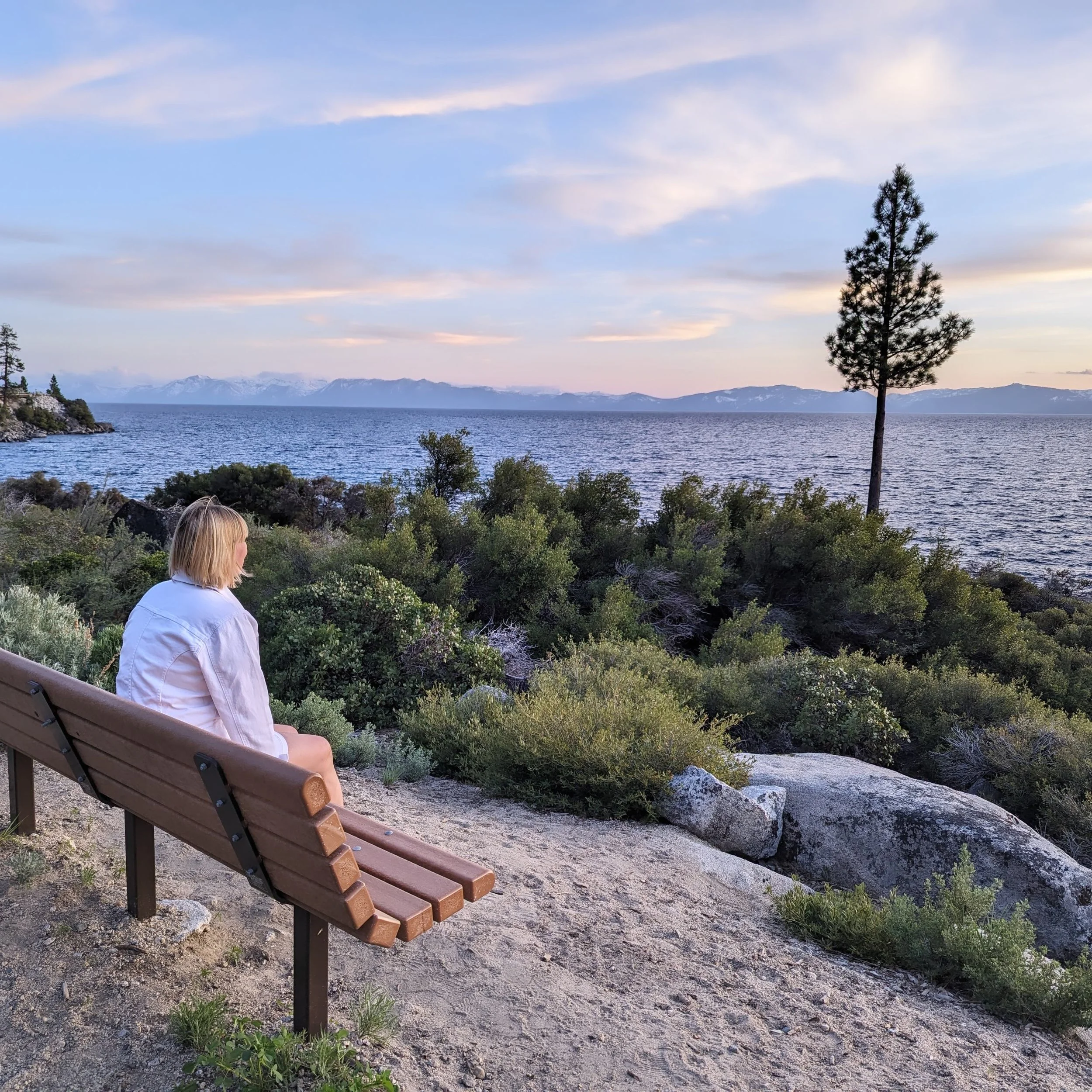 Sally Mayberry sitting on a bench overlooking a peaceful lake and distant mountains at sunset.