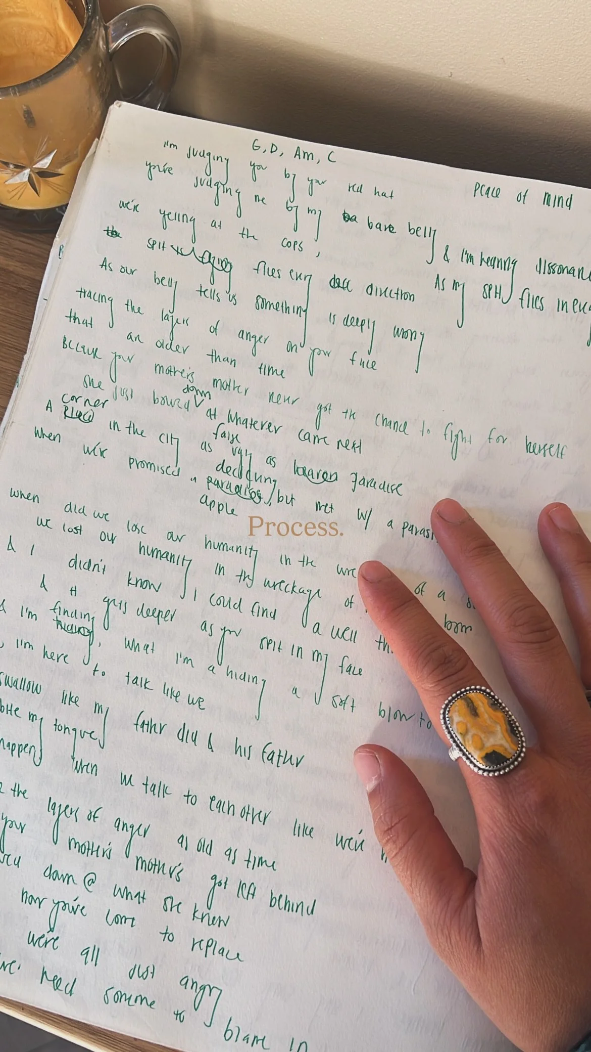A hand with a large decorative ring writes on a white piece of paper with green handwritten text. A candle is visible in the upper left corner of the image.