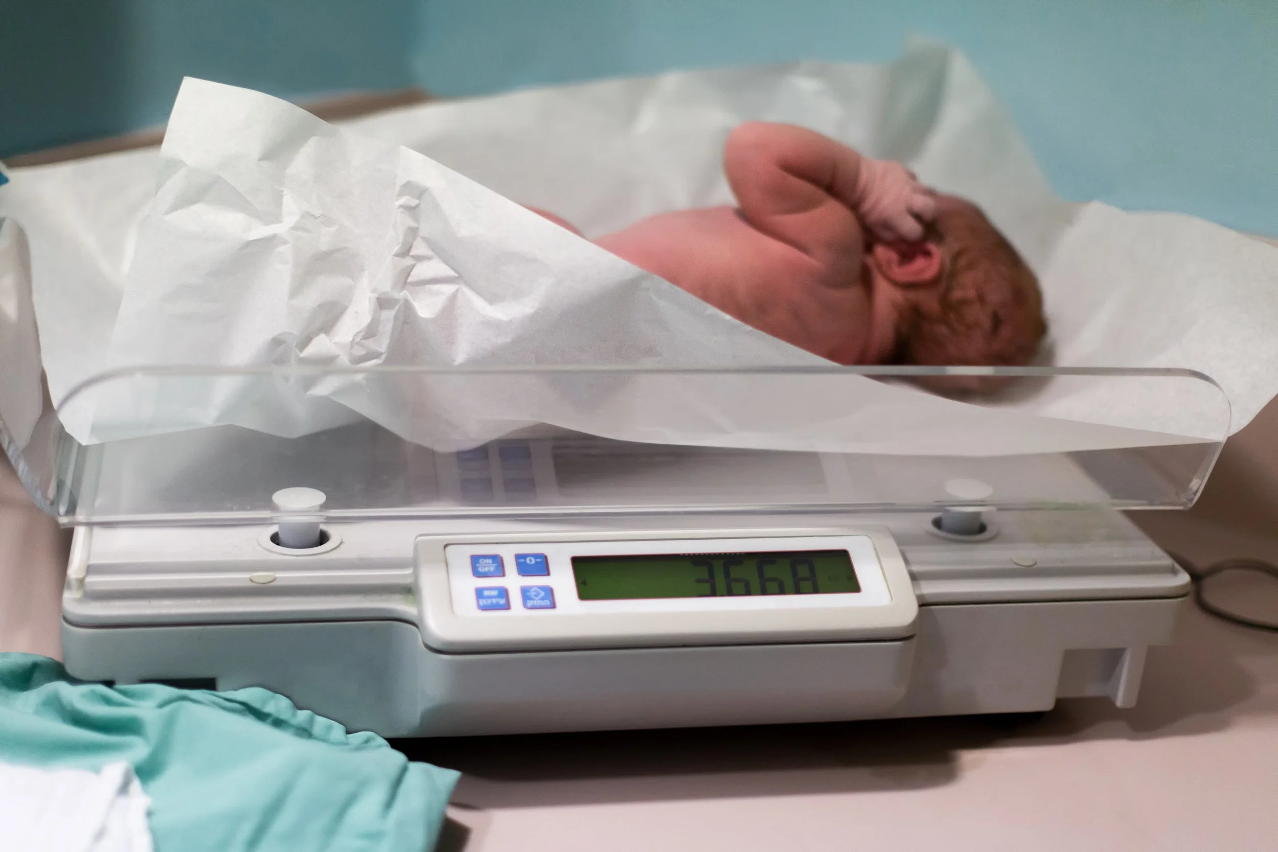 Newborn baby lying on a hospital medical scale being weighed shortly after birth, representing routine newborn weight monitoring in the first days of life
