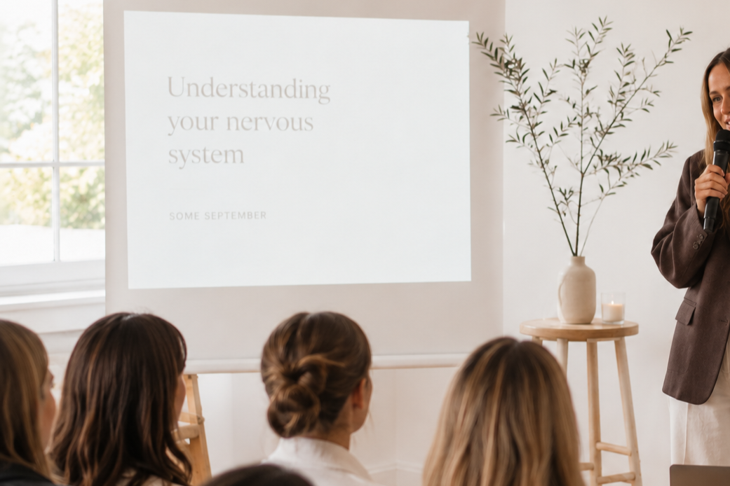 A woman gives a presentation in a room with seated audience members, a slide on NM nervous system is projected on the wall, and a tall plant and candle on a small table are visible.