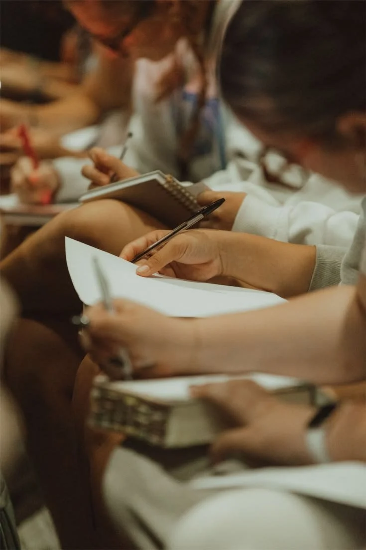 Close-up of multiple people taking notes with notebooks and pens in a meeting or classroom setting.