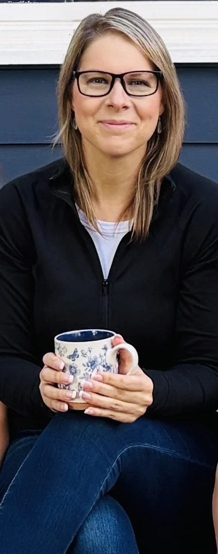 Woman with glasses sitting outside, holding a floral patterned mug, smiling.