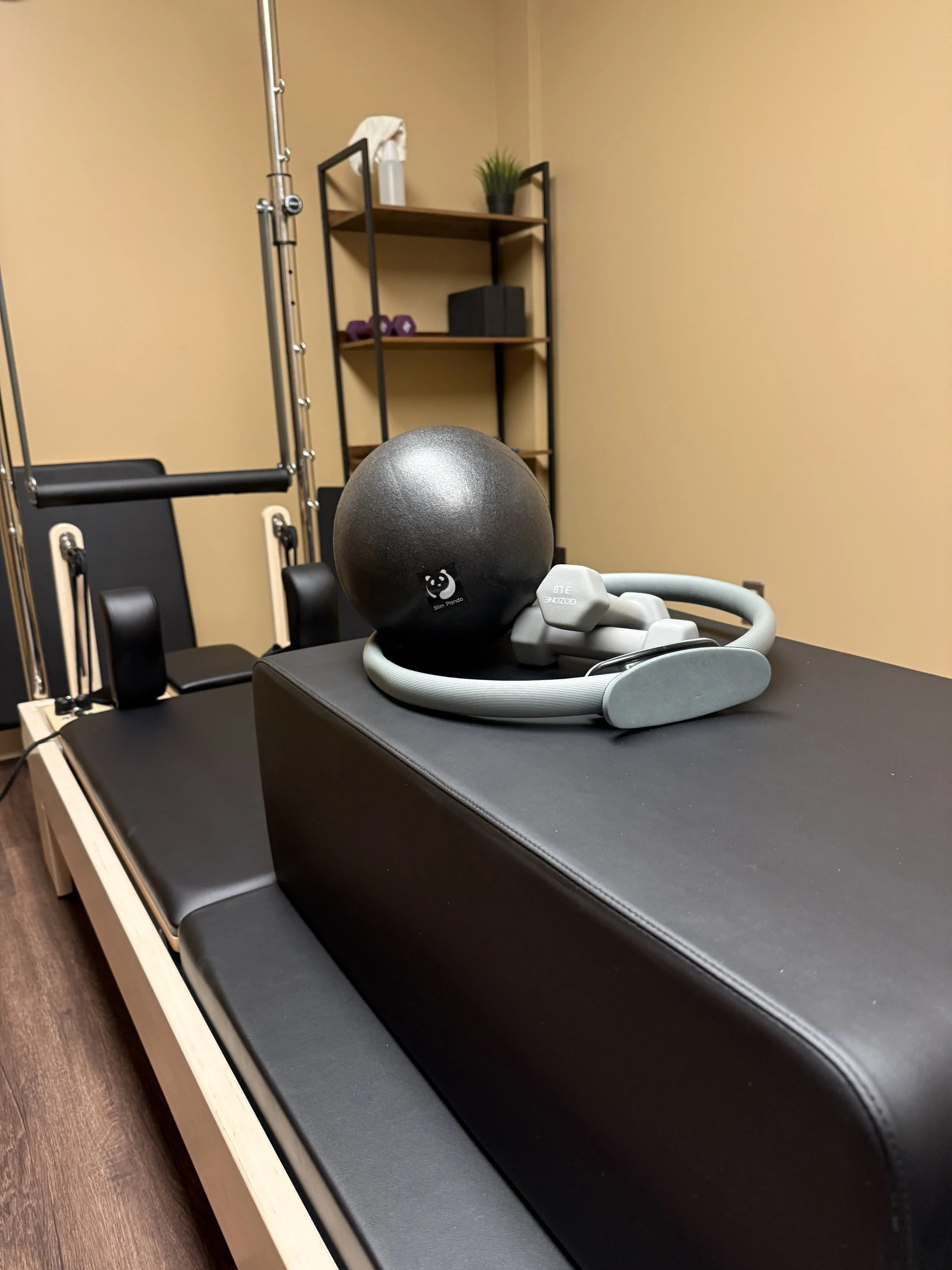 Exercise equipment including a black medicine ball, a pair of gray headphones, and a black massage table in a room with beige walls and a wooden shelf holding a plant, a spray bottle, and small weights.