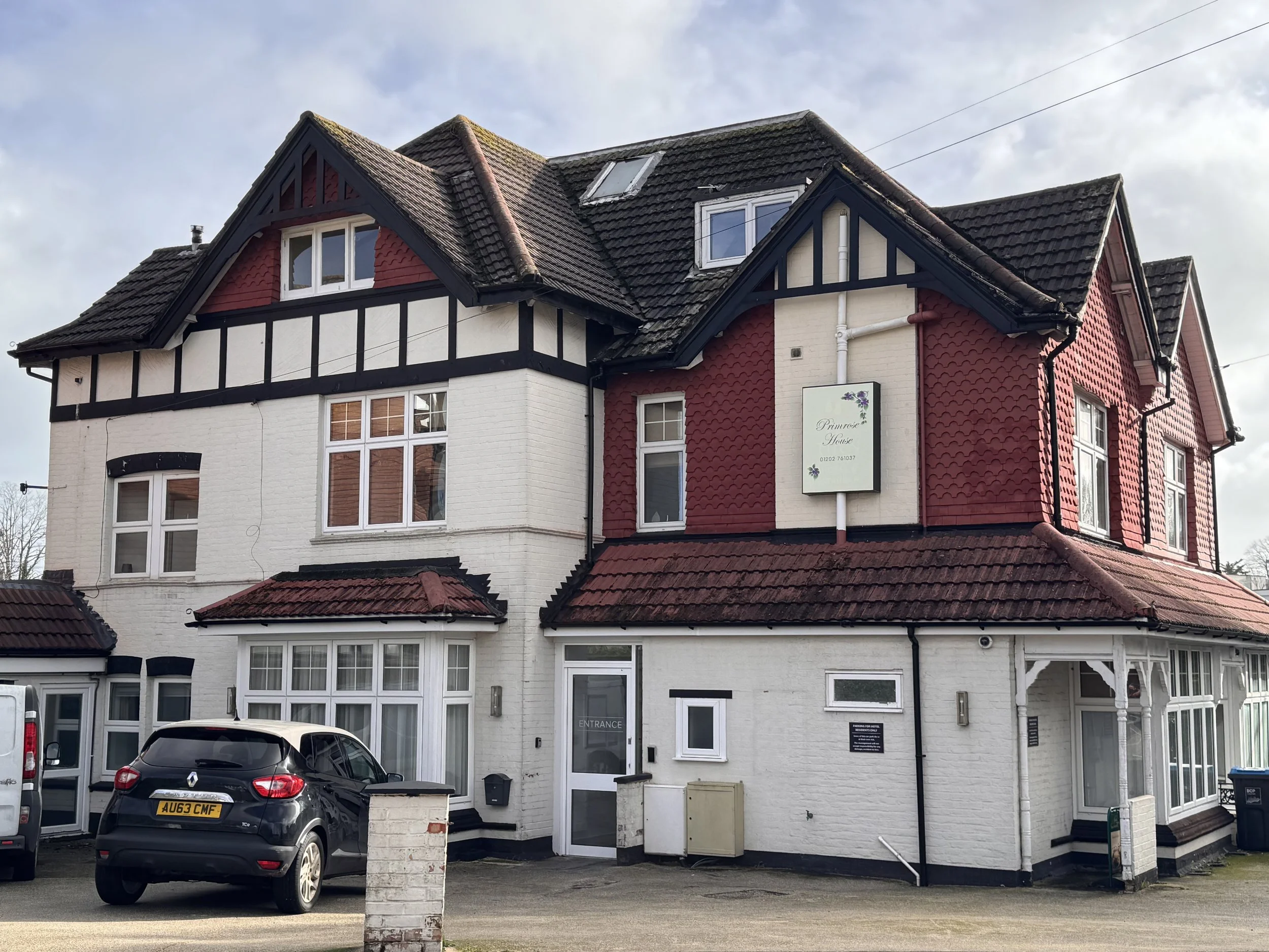 A multi-story building with white and red walls, black framework, and a tiled roof. There are several windows, a parking area with two cars, and a sign on the building.