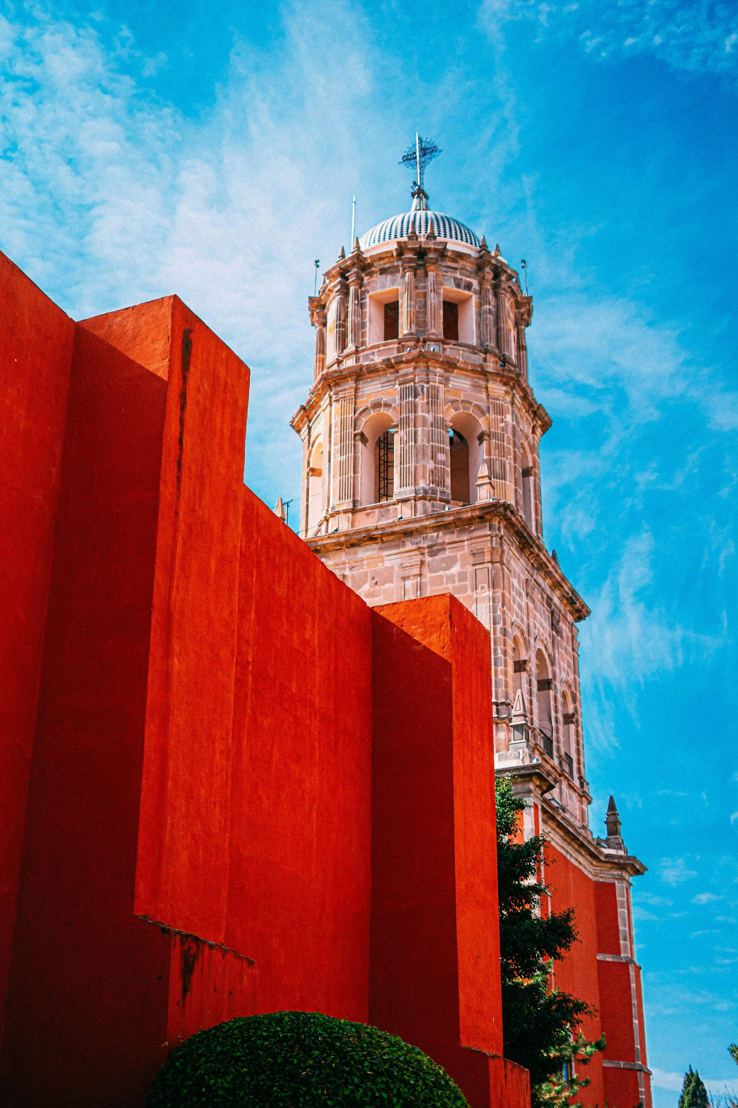 A tall, historic pink stone tower with arched windows and a weather vane on top, against a bright blue sky with wispy clouds. In the foreground, there is a red wall and some greenery at the base.