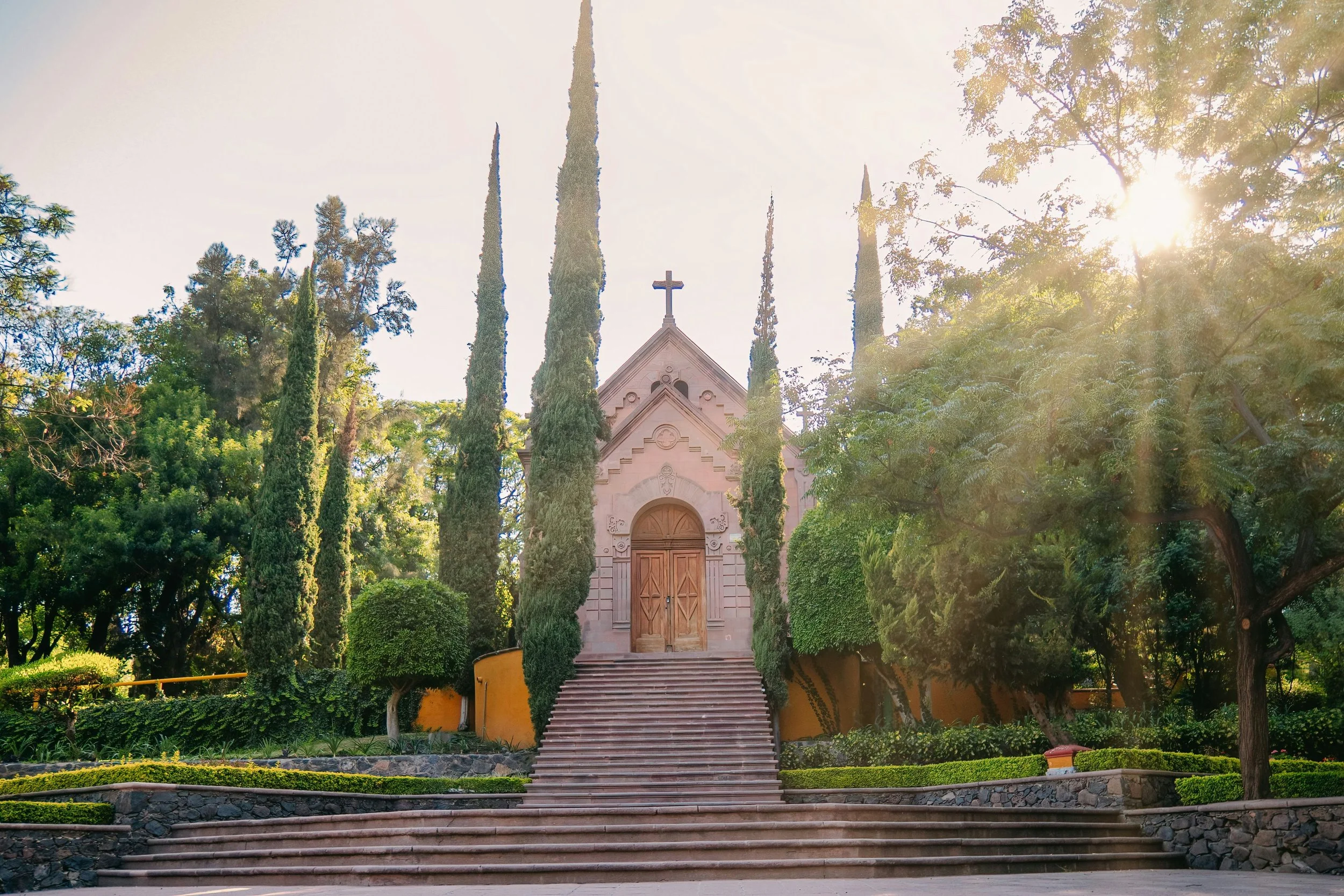A small church with a cross on top, surrounded by tall cypress trees and lush greenery, with sunlight filtering through the trees.
