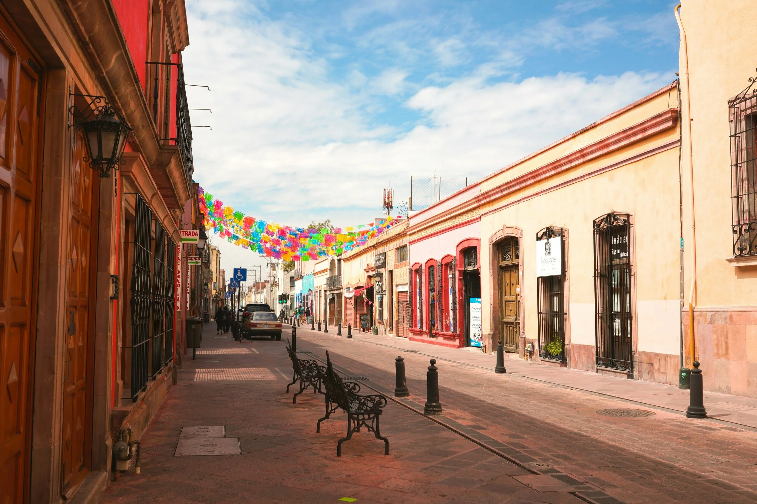 Colorful decorations hang across a quiet street with benches, parked cars, and buildings with ornate windows and doors under a partly cloudy sky.