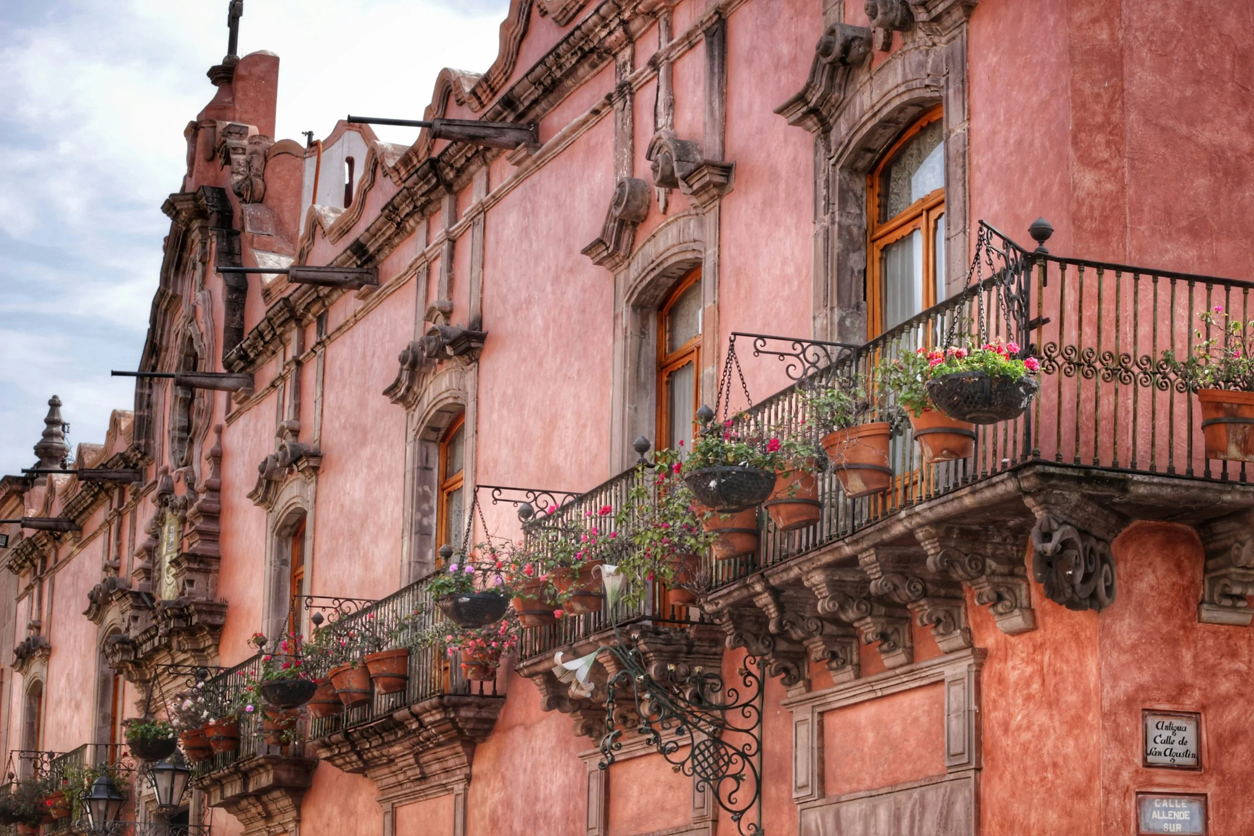 Pink historic building with ornate stone window frames and iron balconies decorated with flower pots filled with colorful flowers.