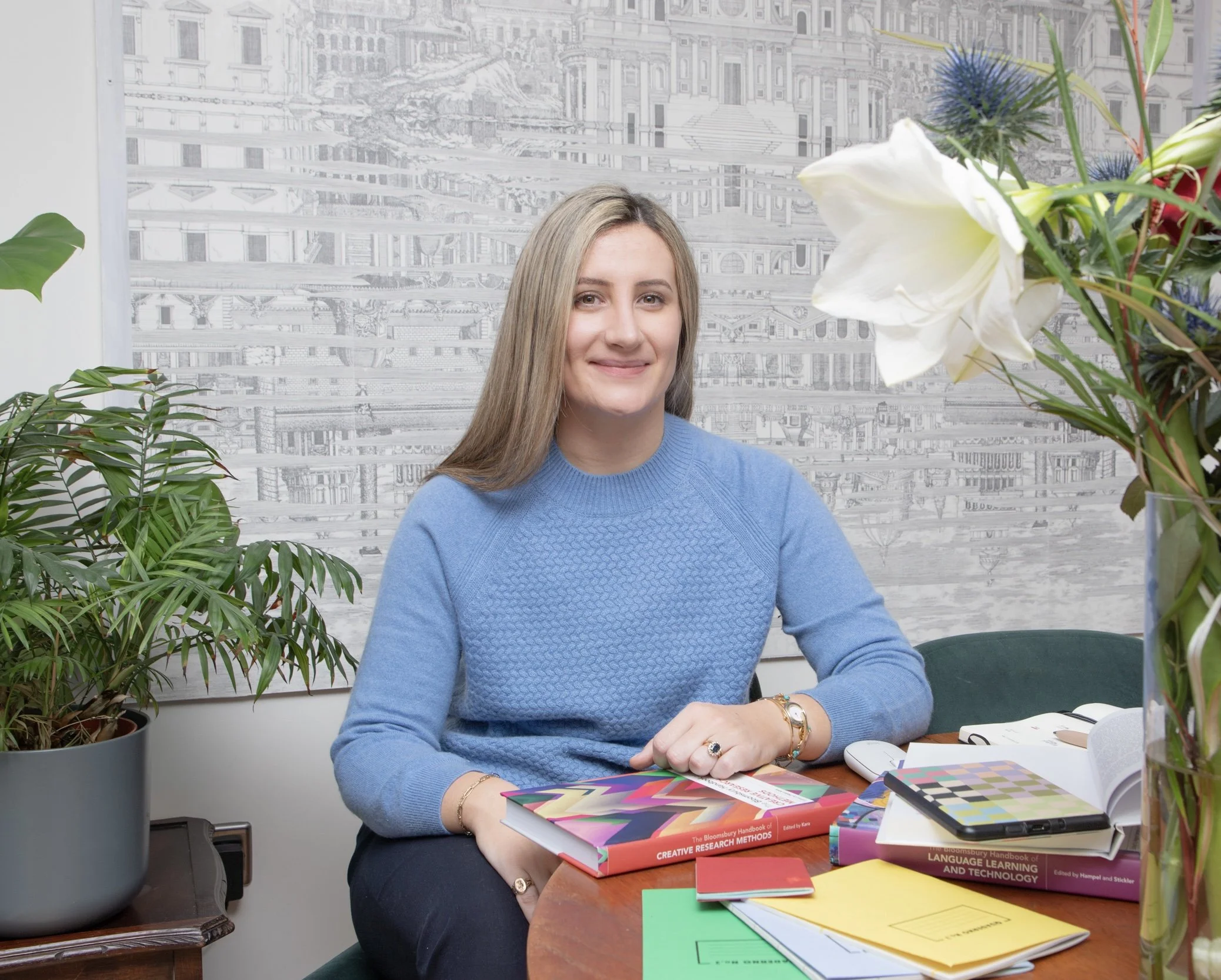 A woman with long blonde hair and fair skin, wearing a light blue sweater, is seated at a desk cluttered with colorful books, notebooks, and a tablet. She is smiling and looking at the camera. There are green plants on her left and a large abstract cityscape framed artwork behind her.
