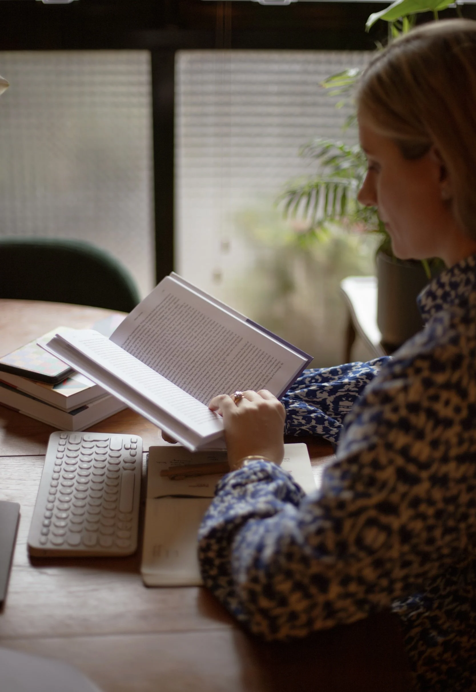 A woman sitting at a desk reading a book, with a smartphone and stacks of books nearby, and a window with blurred outdoor scenery in the background.