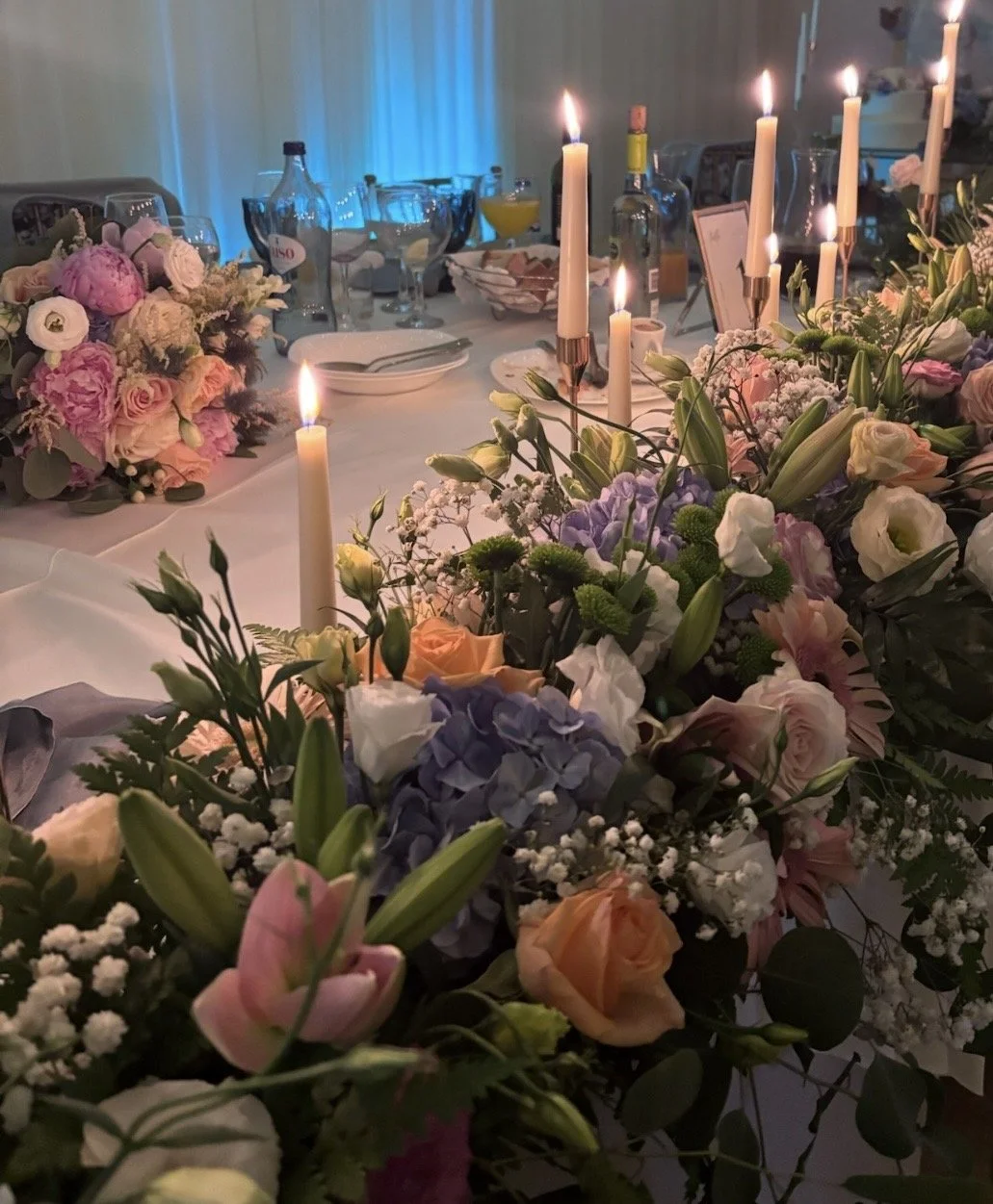 A decorated dining table with floral arrangements, lit candles, plates, glasses, and beverages, set for an elegant event or celebration.