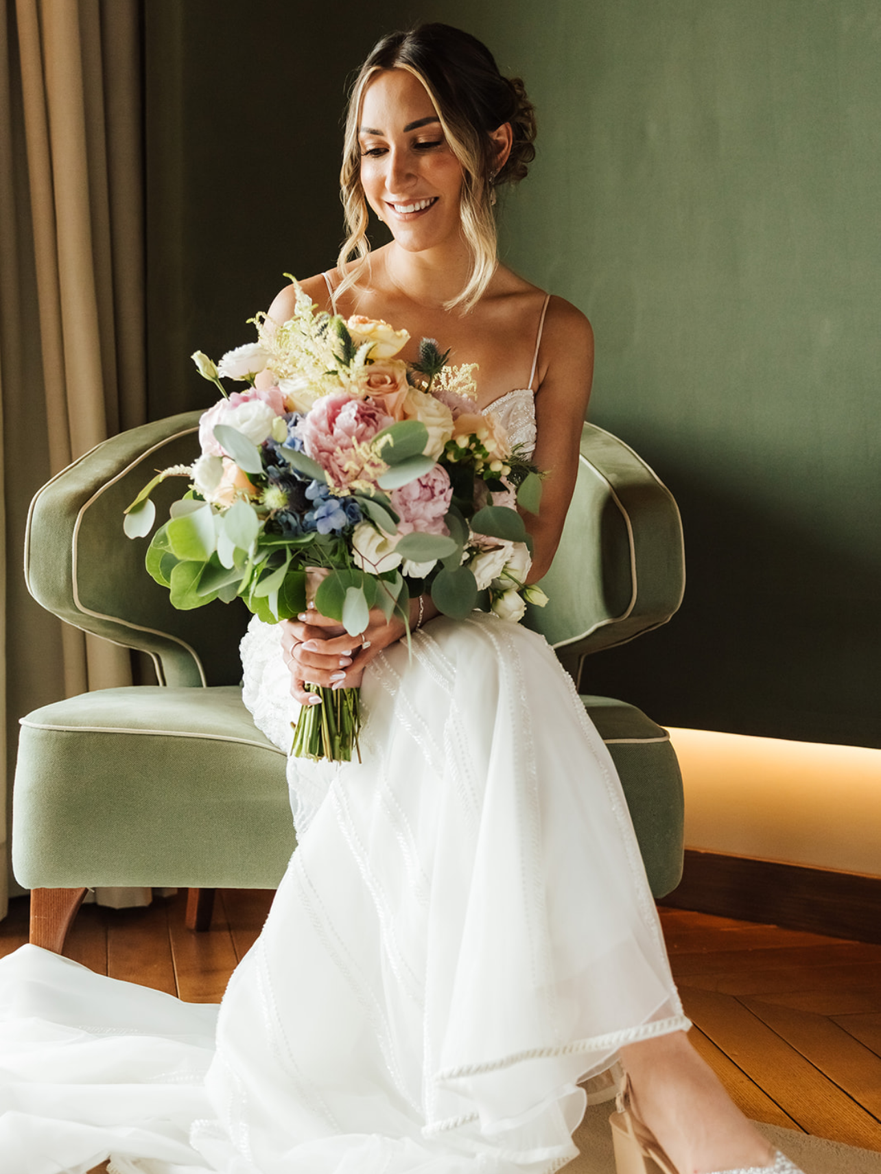 Bride in a white wedding dress sitting on a green chair, holding a large bouquet of flowers, smiling while looking down.