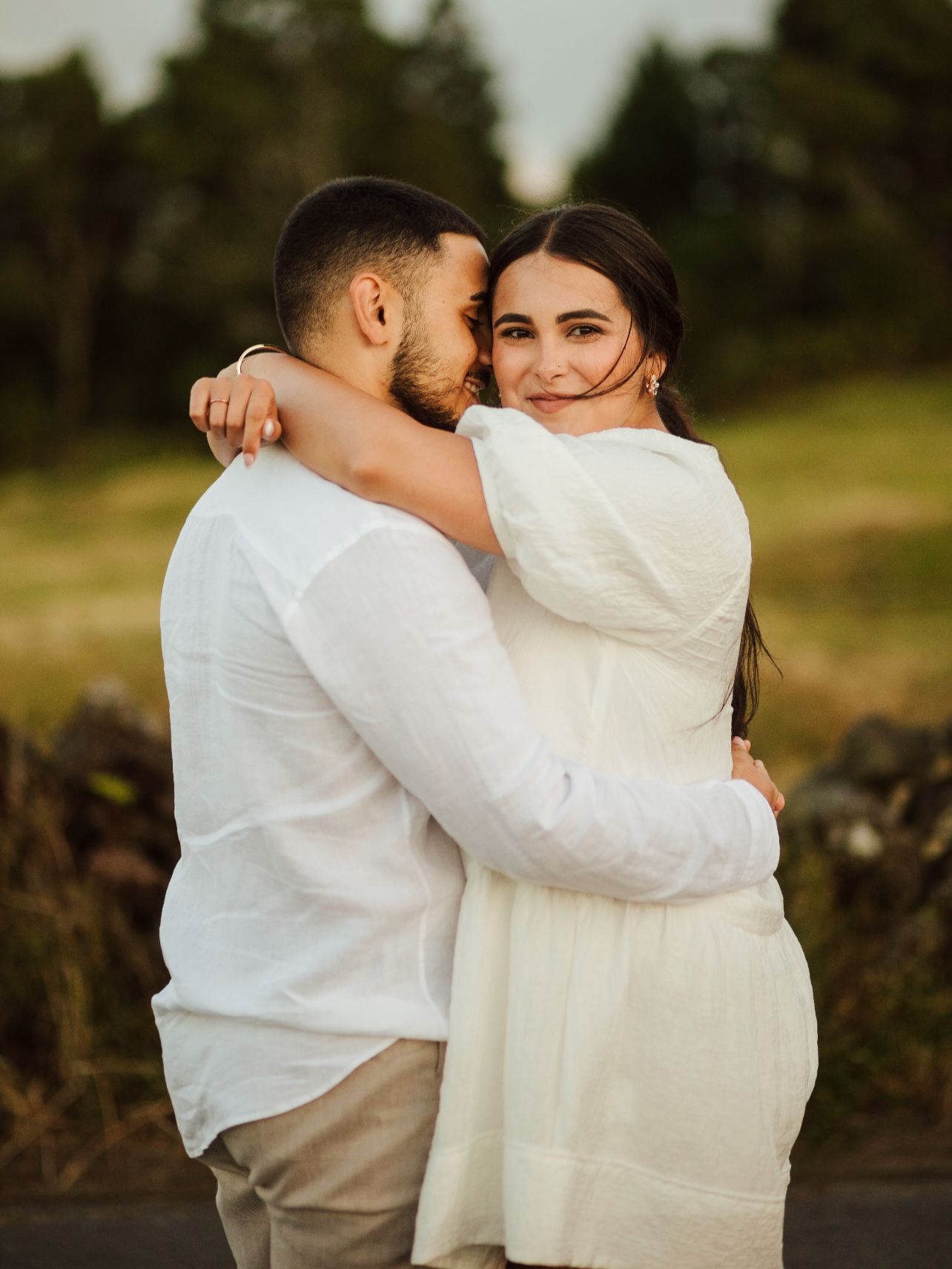 A couple embracing outdoors during sunset, smiling at the camera, with trees in the background.