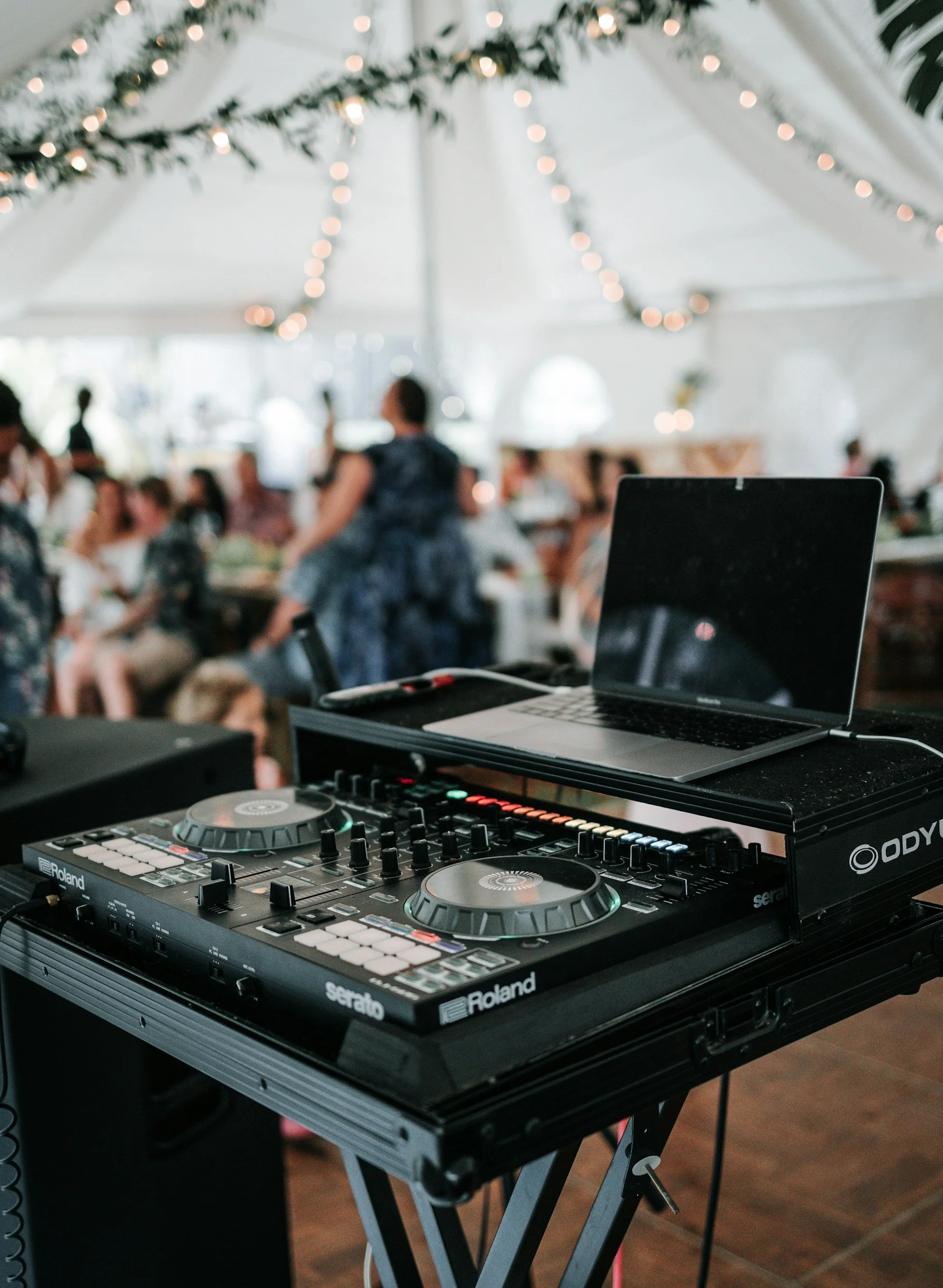 A DJ setup with a Roland DJ controller and a laptop on a stand, with a blurred background of people at an event decorated with string lights and greenery.