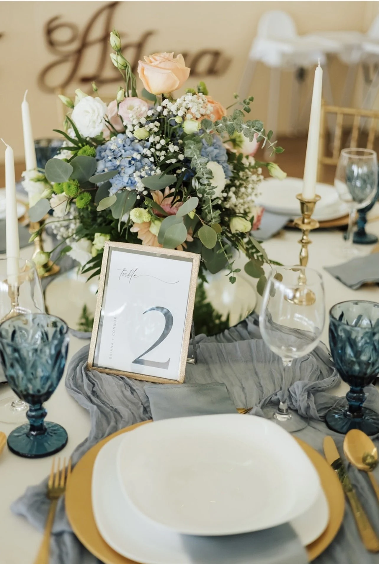 Elegant table setting with a floral centerpiece featuring roses, hydrangeas, and eucalyptus, surrounded by tall white candles in gold holders, blue glassware, a white and gold plate, gold cutlery, and a table number sign with the number 2.