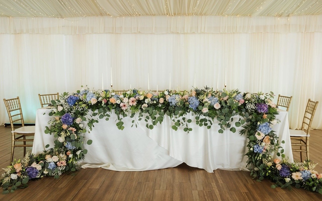 Elegant wedding reception table decorated with a floral arrangement of pink, purple, and white flowers and greenery, set against a backdrop of white drapes with string lights.