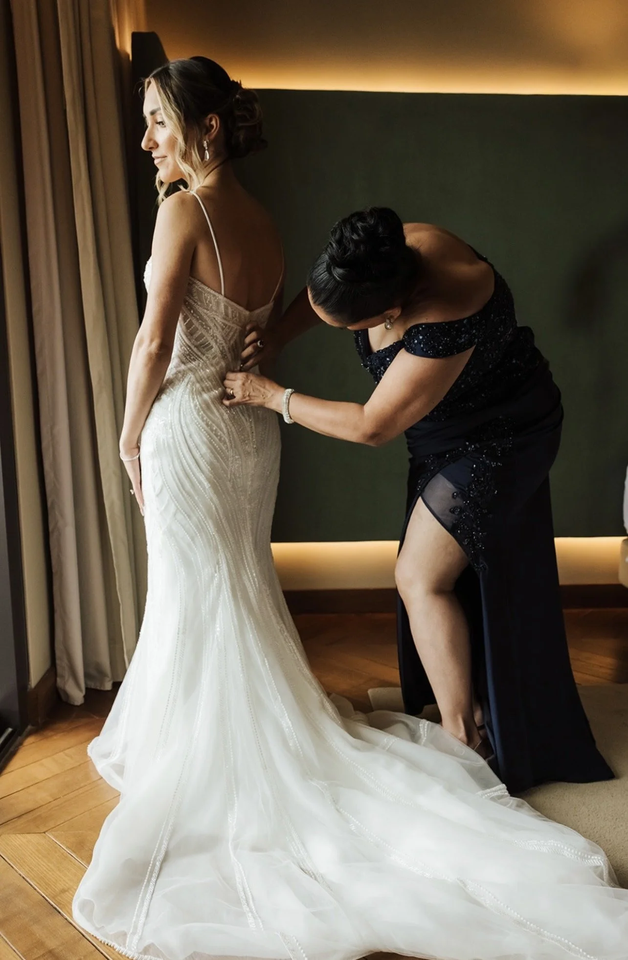 A bride in a white wedding gown with spaghetti straps is being helped with her dress by a woman in a black dress. The bride is standing near a window, and the gown has detailed beadwork and a long train. The woman adjusting the dress is leaning over,