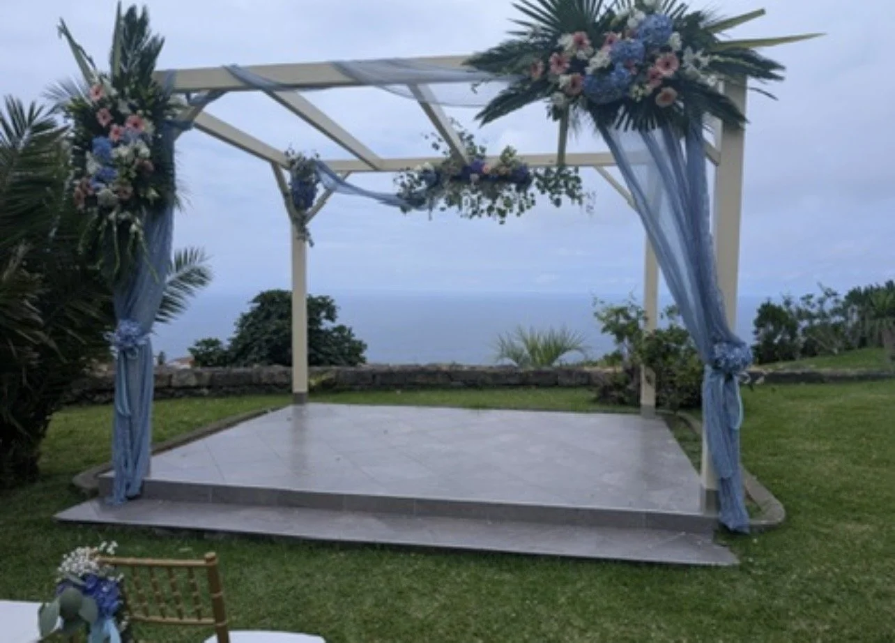 An outdoor wedding arch decorated with pink, blue, and white flowers, and blue drapes, on a tiled platform overlooking a body of water and surrounded by lush green grass and plants.