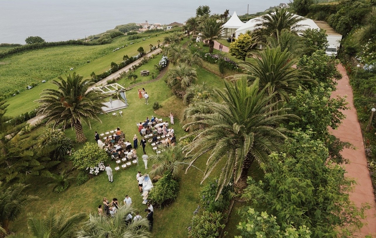 Outdoor wedding ceremony setup in a lush garden with palm trees, chairs arranged for guests, and a small stage with performers, overlooking a body of water in the distance.