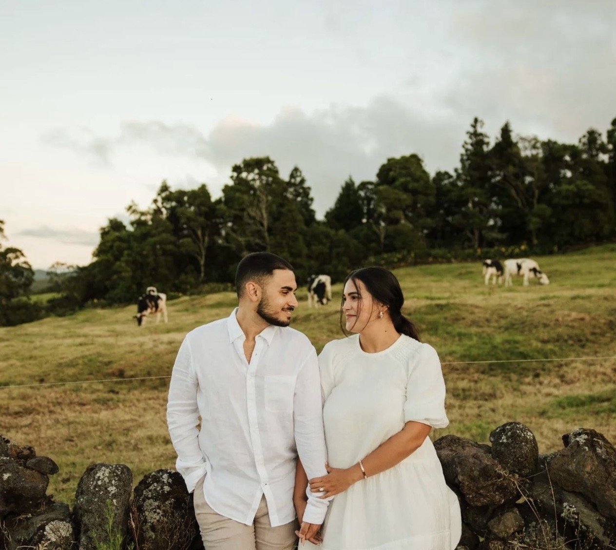 A couple stands close together, holding hands, outdoors in a rural setting with cows grazing in the background and trees on a cloudy evening.